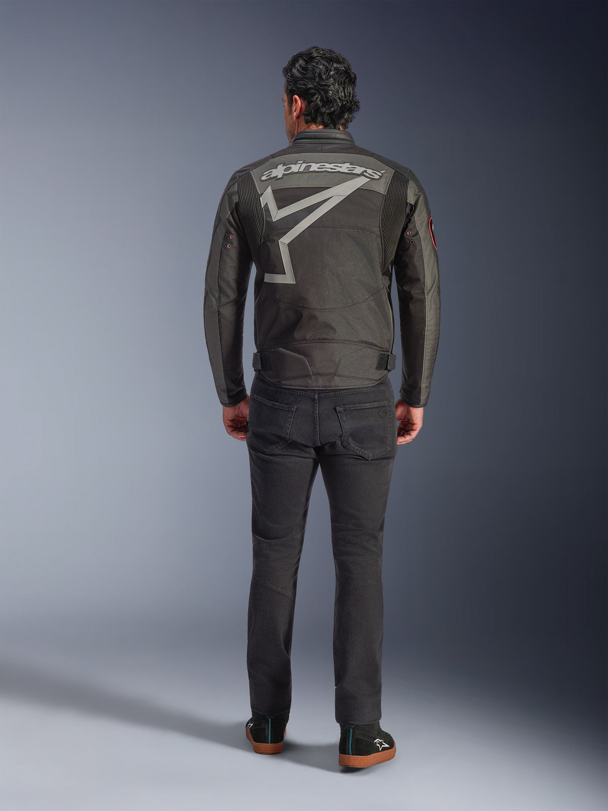 Alpinestars Axton Jacket, Motorcycle Jacket, Black Anthracite Red Tangerine, back view showing large grey Alpinestars logo and star graphic, accordion shoulder panels, worn with black pants and casual black shoes.
