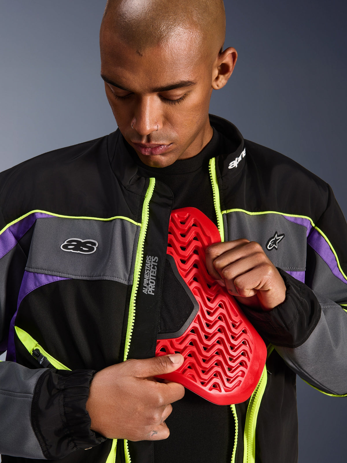 Alpinestars Unite Jacket, Urban Motorcycle Jacket, Black Asphalt and Violet with neon yellow accents, male model demonstrating a red Nucleon chest armor insert inside the open jacket, minimalist streetwear aesthetic for urban riding.