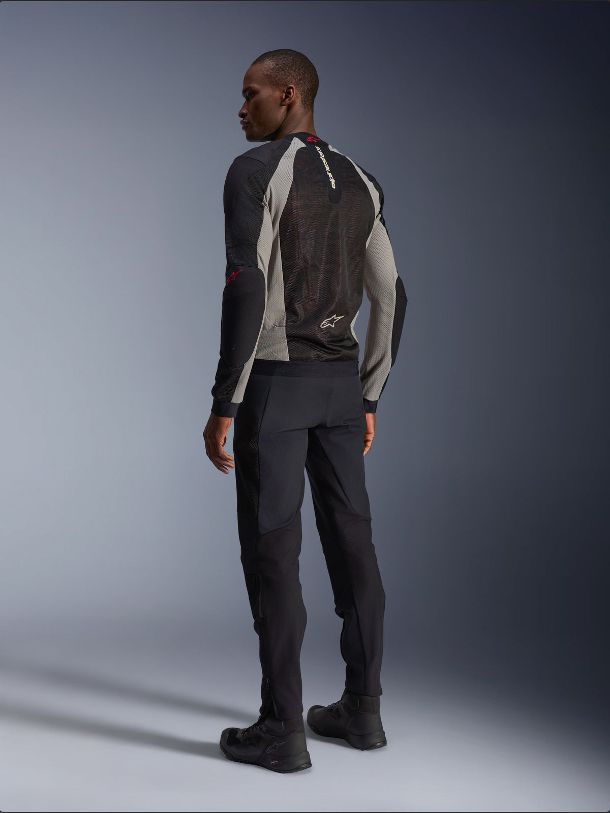 Model wearing Alpinestars Troop-Air Jacket from the back, Black Ash Gray and Bright Red accents, technical mesh construction for ventilation, paired with black motorcycle pants and riding boots, urban contemporary style