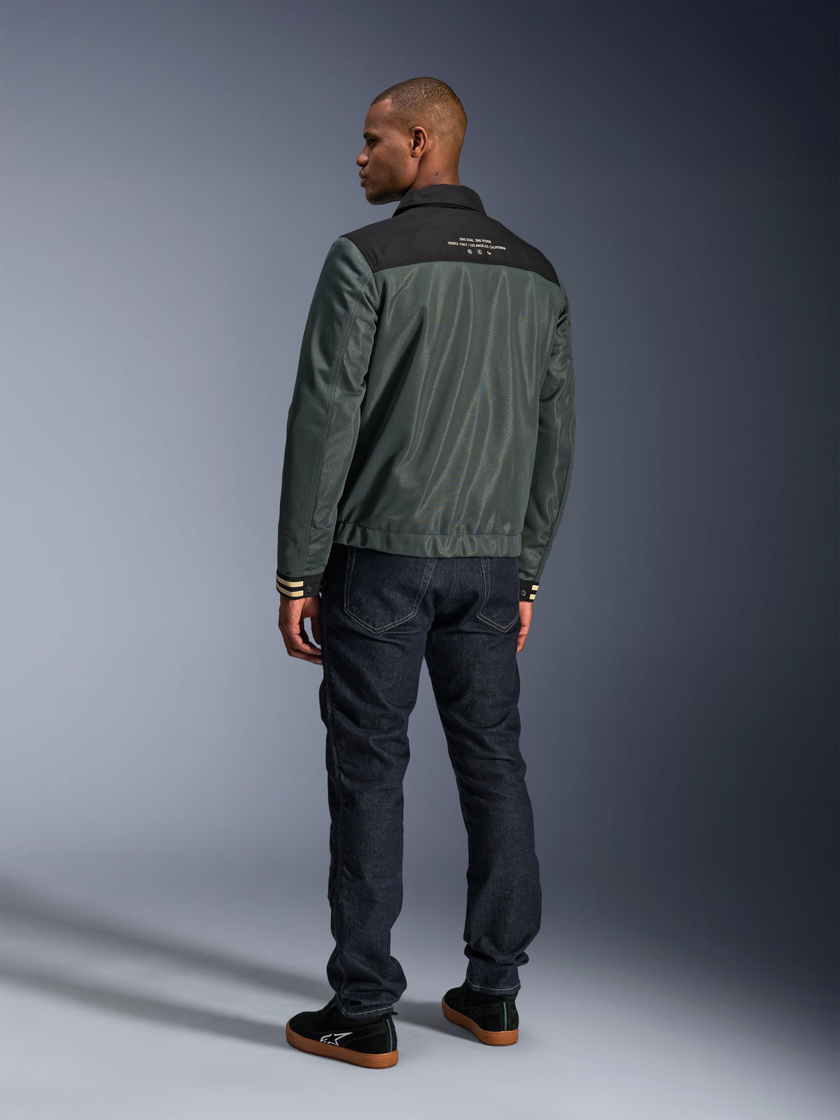 Alpinestars Ignite Air Jacket in Urban Gray Black Blue, rear view on model, featuring a mesh body with a black shoulder yoke and white text, striped cuffs, paired with dark denim jeans and black sneakers, urban lifestyle motorcycle apparel