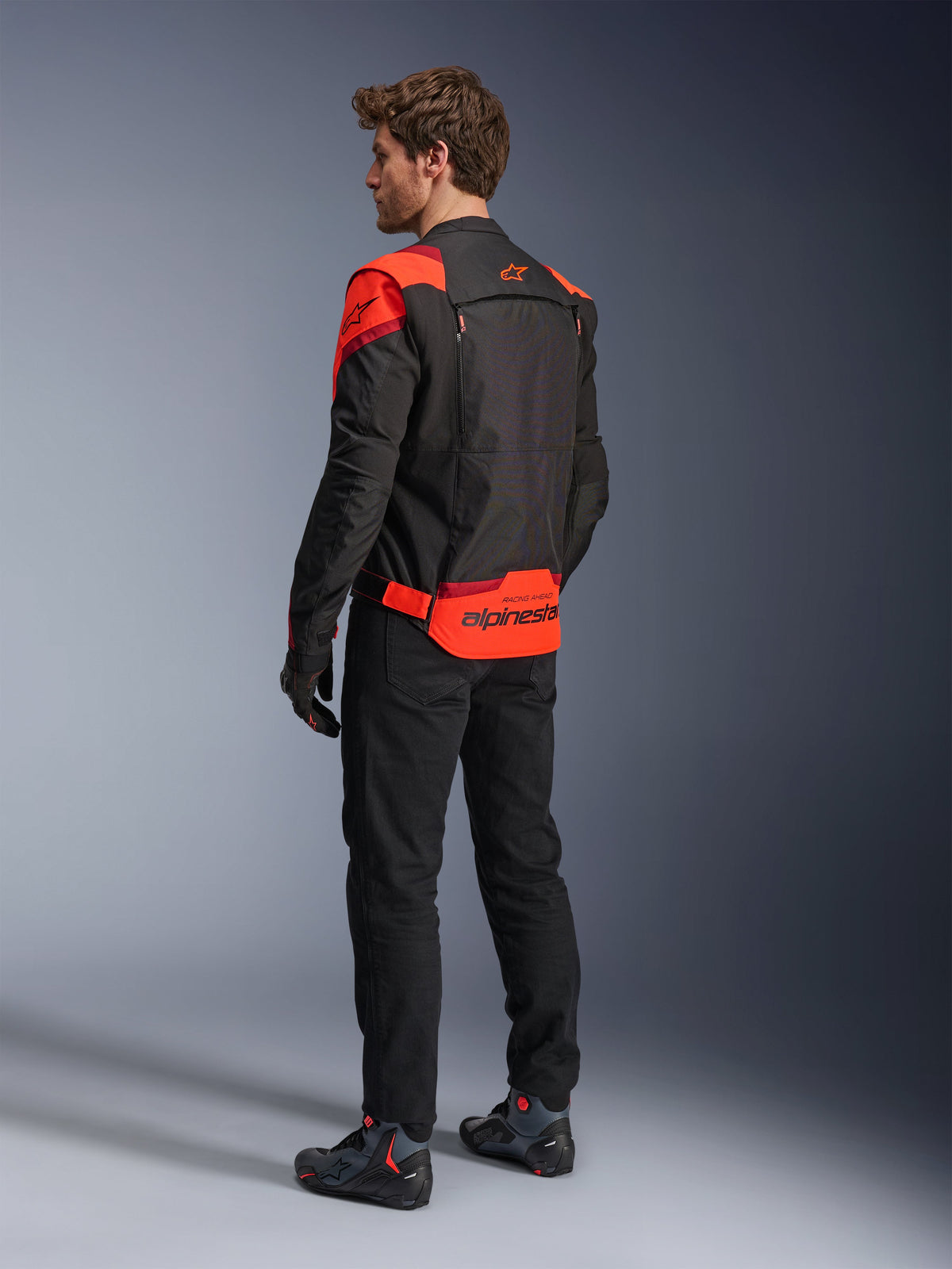 Man wearing Alpinestars T-Stunt Air Jacket from the back, Black Red Fluo and Dark Red, textile motorcycle jacket with mesh panels and sport-cut design, featuring bold Alpinestars branding on the lower back, styled with black pants and riding boots