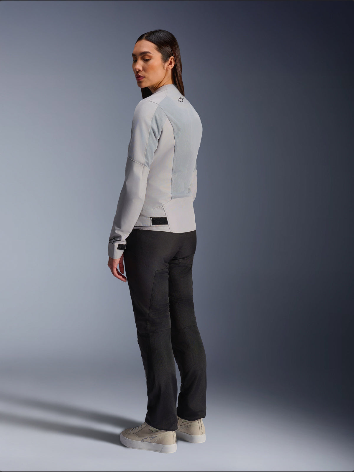 Woman wearing Alpinestars Stella C-1 Air Jacket in Silver, back and side view showing tech mesh panels and reflective shoulder logo, paired with black motorcycle pants and beige sneakers, contemporary urban riding aesthetic