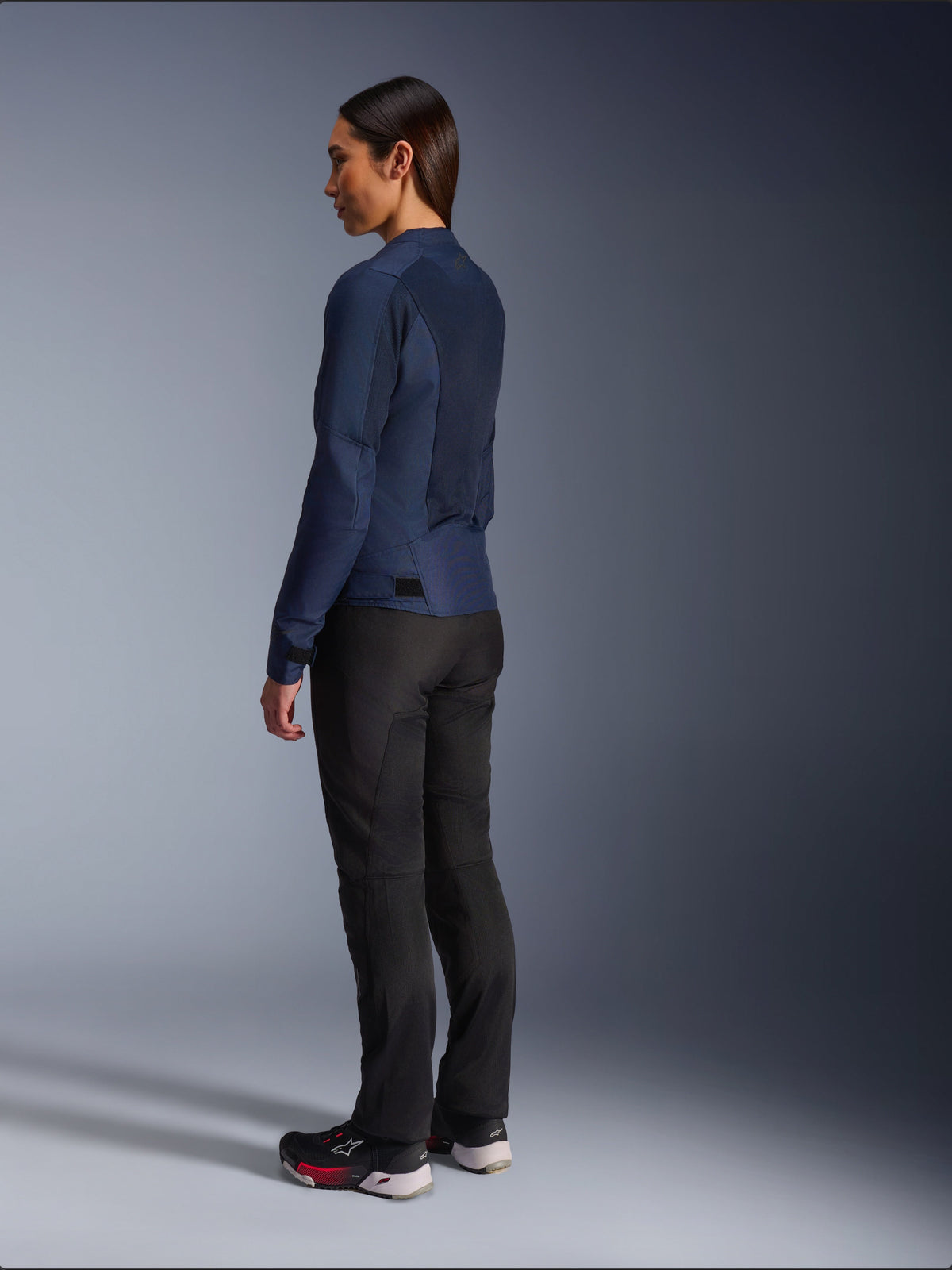 Woman wearing Alpinestars Stella C-1 Air Jacket in Blue, rear three-quarter view, showcasing mesh back panels and adjustable waist tabs, paired with black riding pants and black sneakers, urban motorcycle apparel