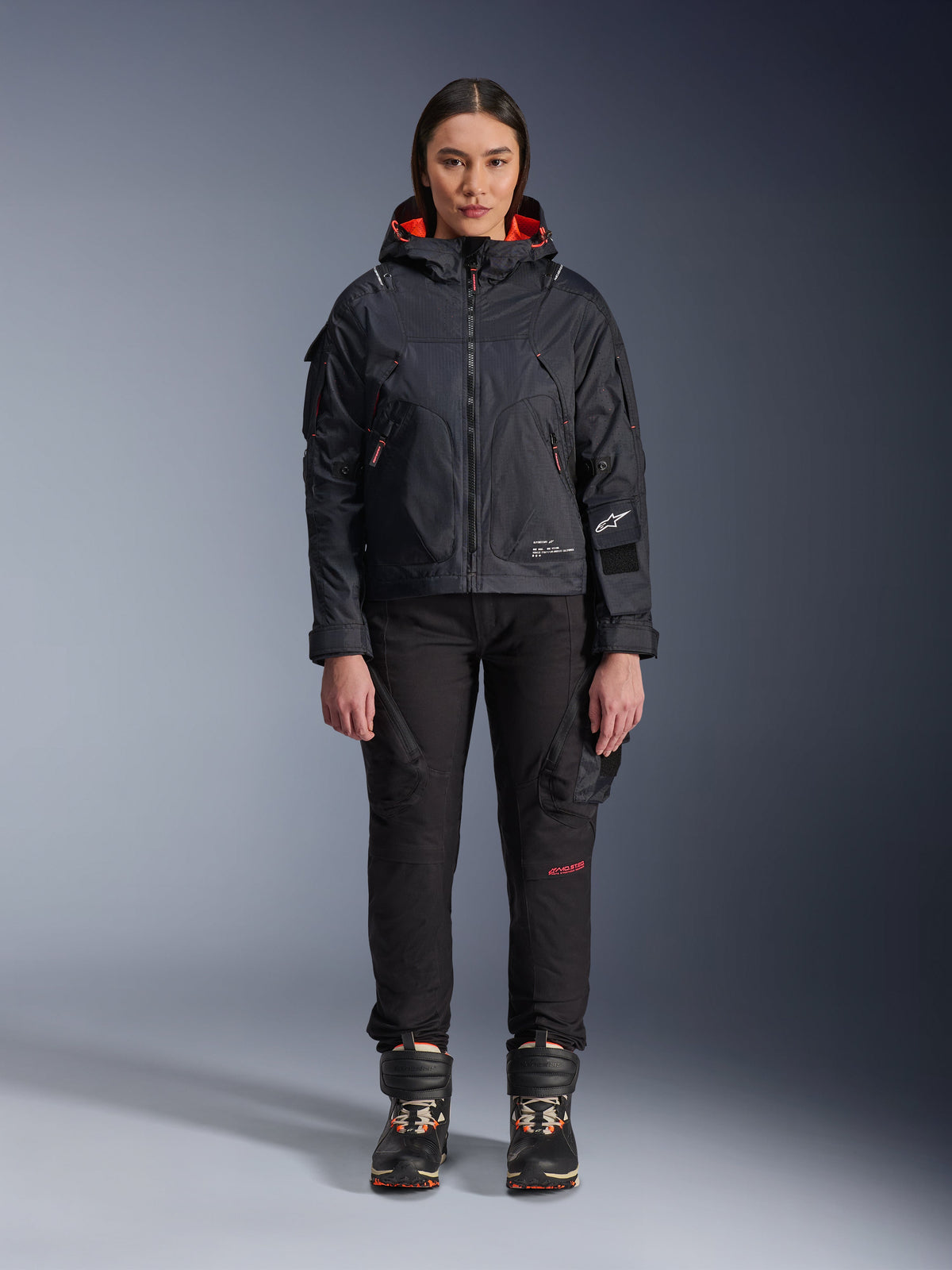 Female model wearing the Alpinestars Molly Air Jacket in Black and Fluo Coral, featuring a utility-inspired design with a hood and coral inner lining, paired with black riding pants and motorcycle boots, urban contemporary motorcycle gear