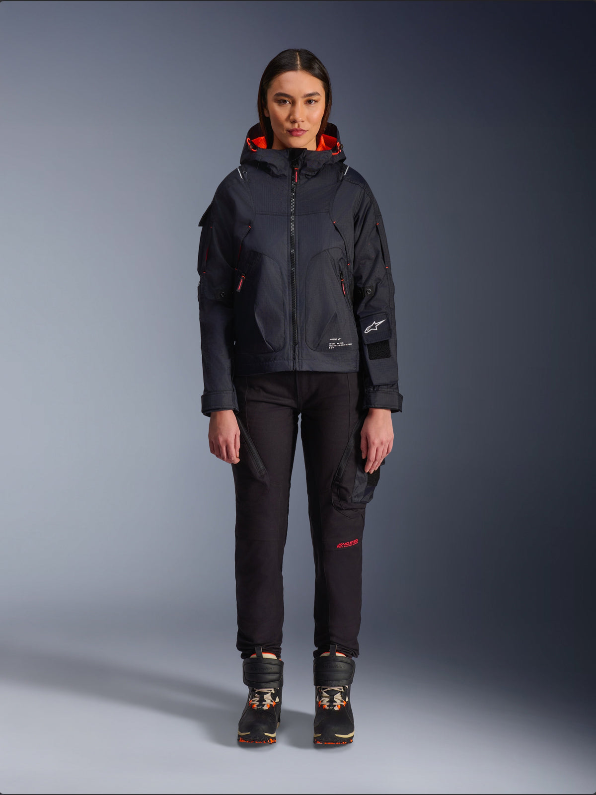 Alpinestars Actea Women's Pants, black motorcycle pants with red text detail and cargo pockets, worn by a female rider with a black hooded jacket, modern urban motorcycle apparel
