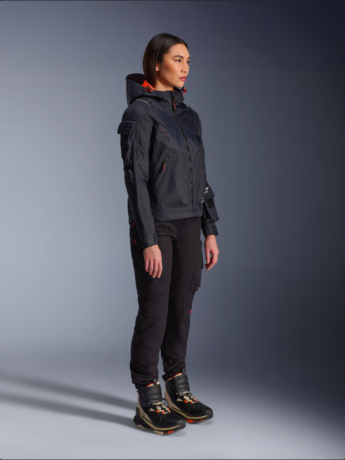Female rider wearing the Alpinestars Molly Air Jacket in Black Fluo Coral, lightweight textile motorcycle jacket with a bright orange hood lining, styled with black riding pants and technical boots, urban contemporary utility-inspired design.