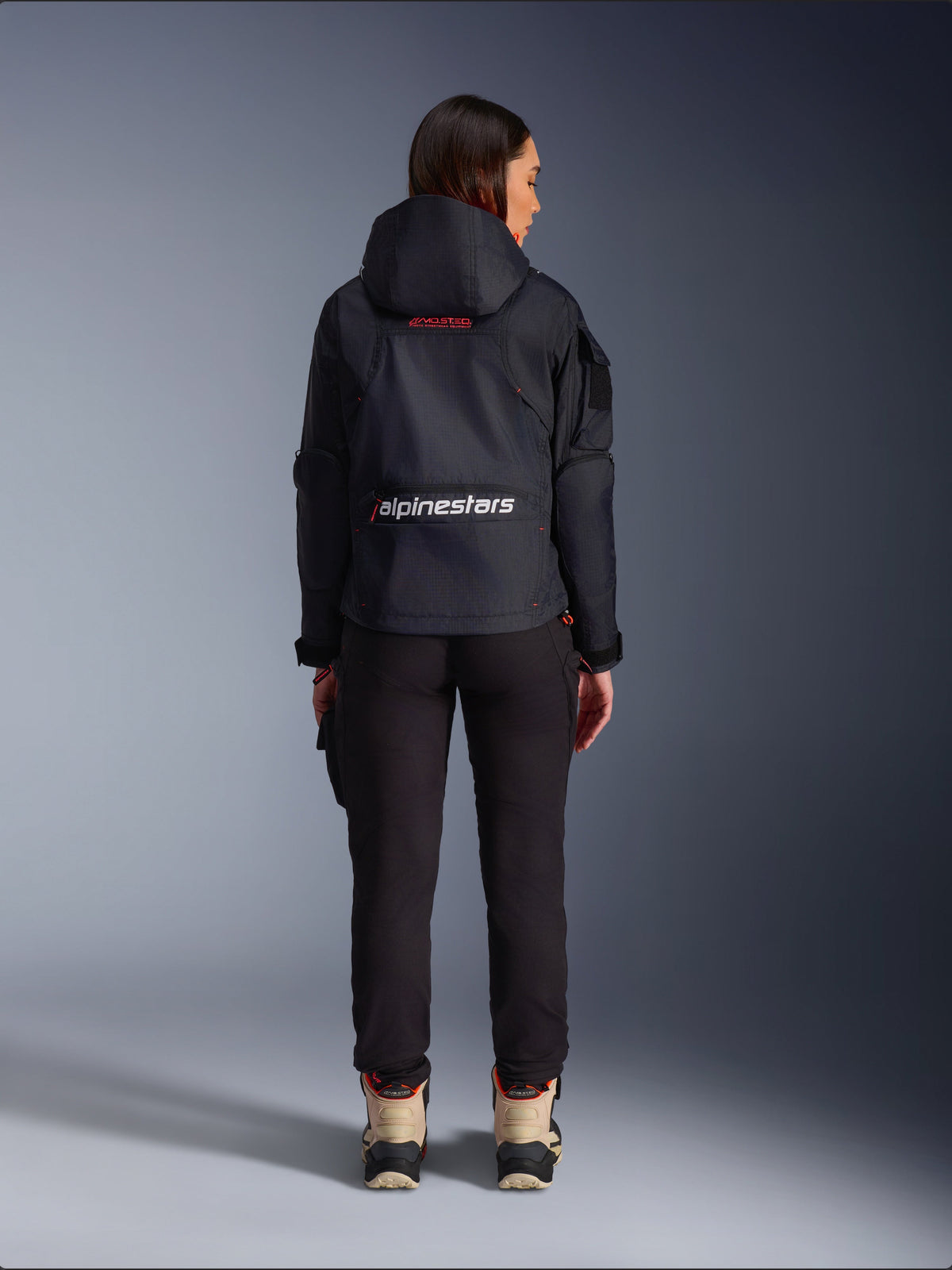 Alpinestars Actea Women's Pants, Black motorcycle riding pants with a tapered fit, paired with a dark hooded Molly Air Jacket and tan boots, rear view of a female rider in urban motorcycling gear