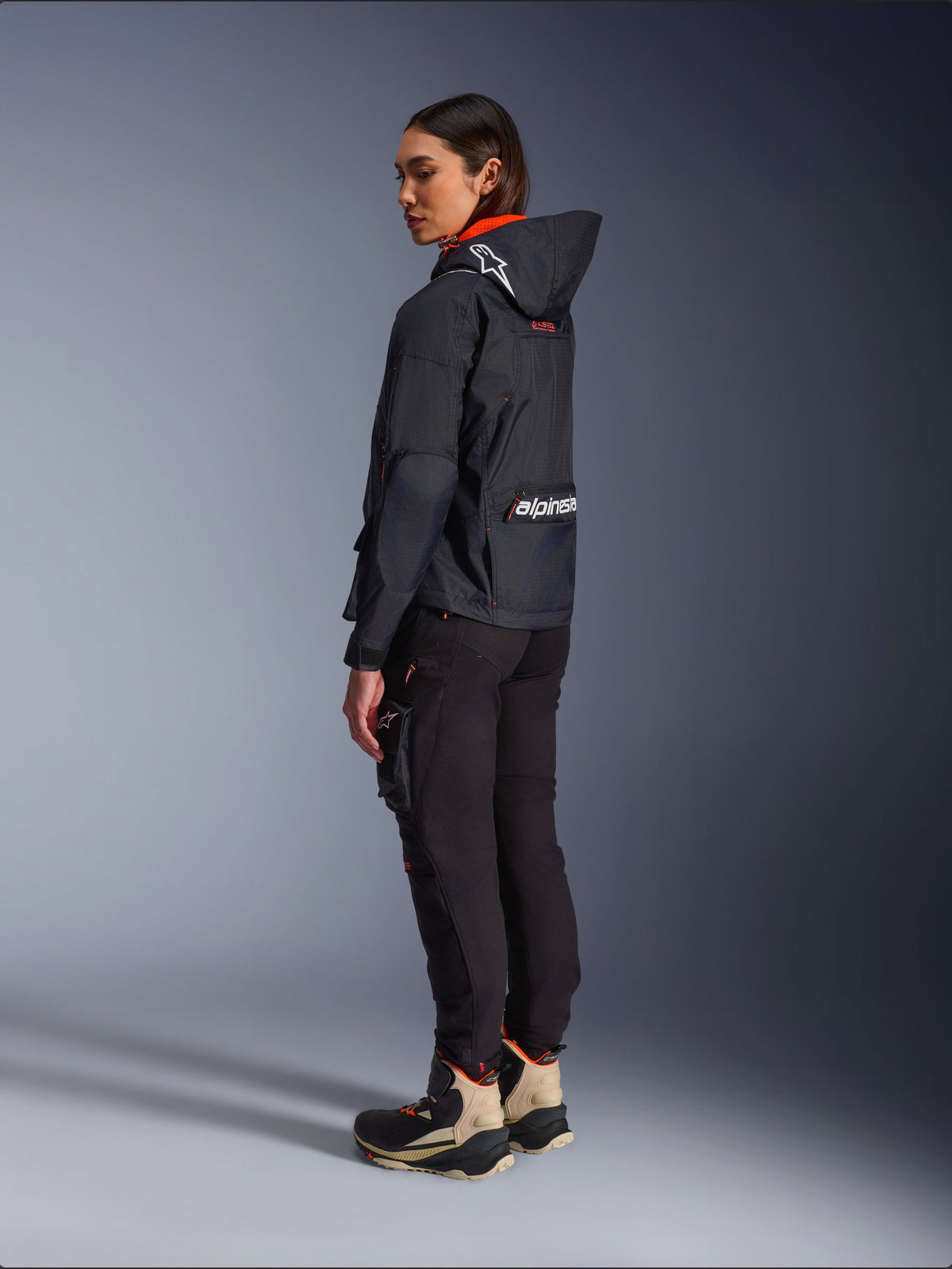 Alpinestars Actea Women's Pants, Black, modern motorcycle pants with a tapered fit and thigh cargo pocket, shown from a side-rear perspective on a model wearing a dark hooded jacket and riding boots, urban riding style.