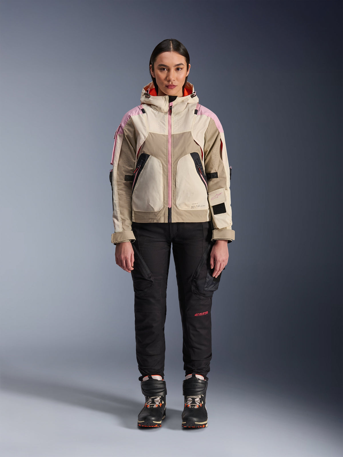 Female model wearing the Alpinestars Molly Air Jacket in Sand Pink Almond, paired with black technical riding pants and boots, front full-body view, urban motorcycle jacket with utility-inspired design and pink accents