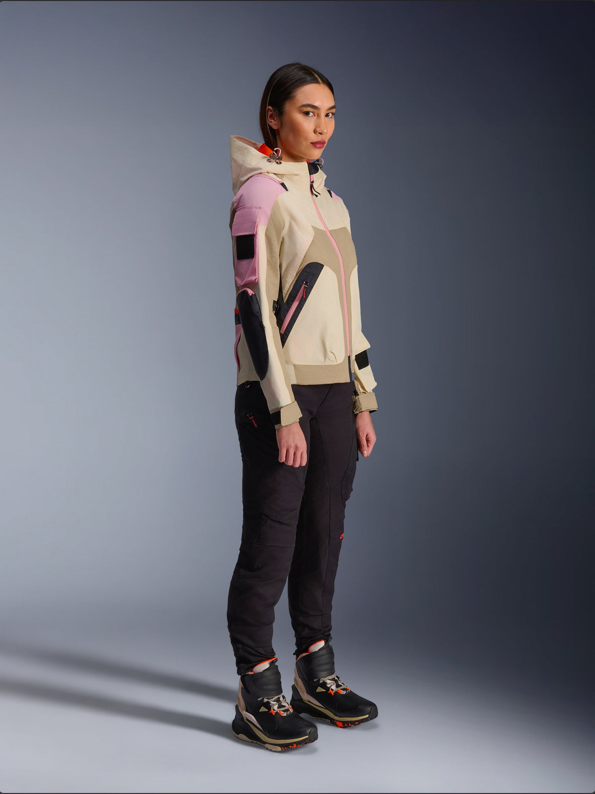 Female model wearing the Alpinestars Molly Air Jacket in Sand Pink Almond, paired with dark technical pants and high-top riding boots, urban motorcycle jacket featuring a hood and multi-color paneling, contemporary streetwear-inspired design