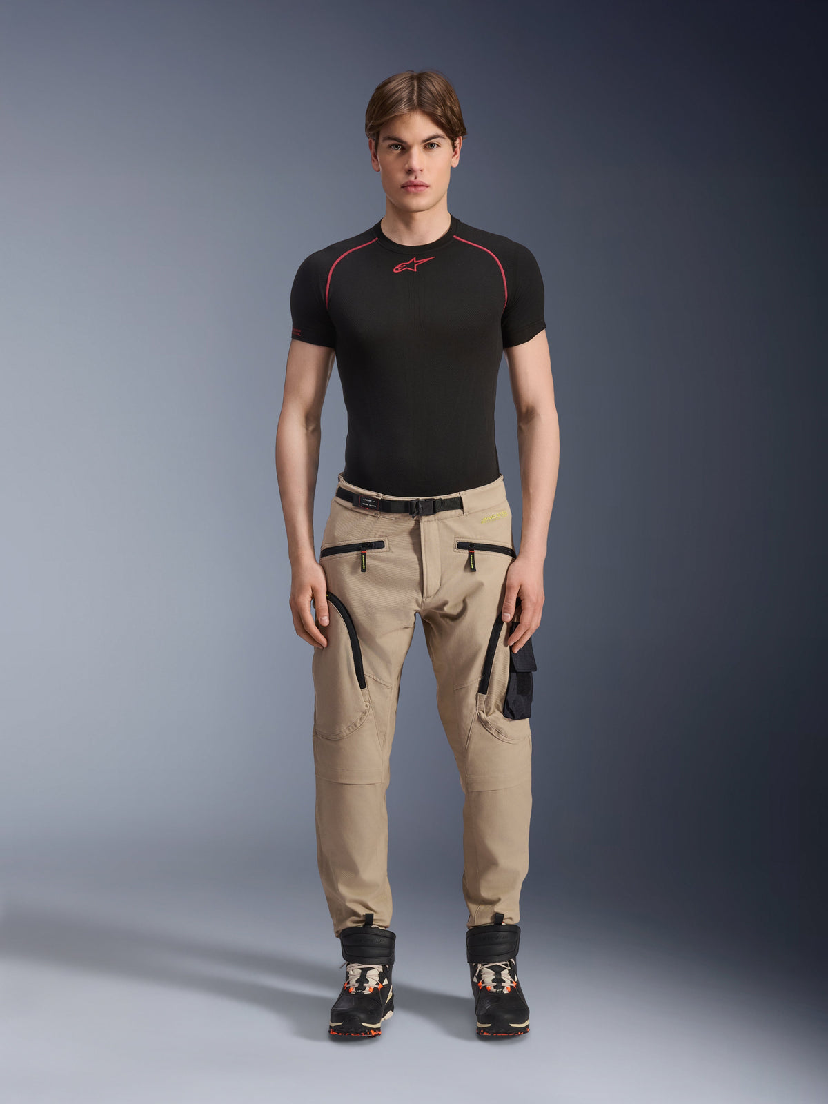 Male model wearing Alpinestars Acteon Pants in Sand, paired with a black short-sleeve base layer and black technical boots, featuring cargo pockets with black zippers and a tapered fit, contemporary urban motorcycle gear