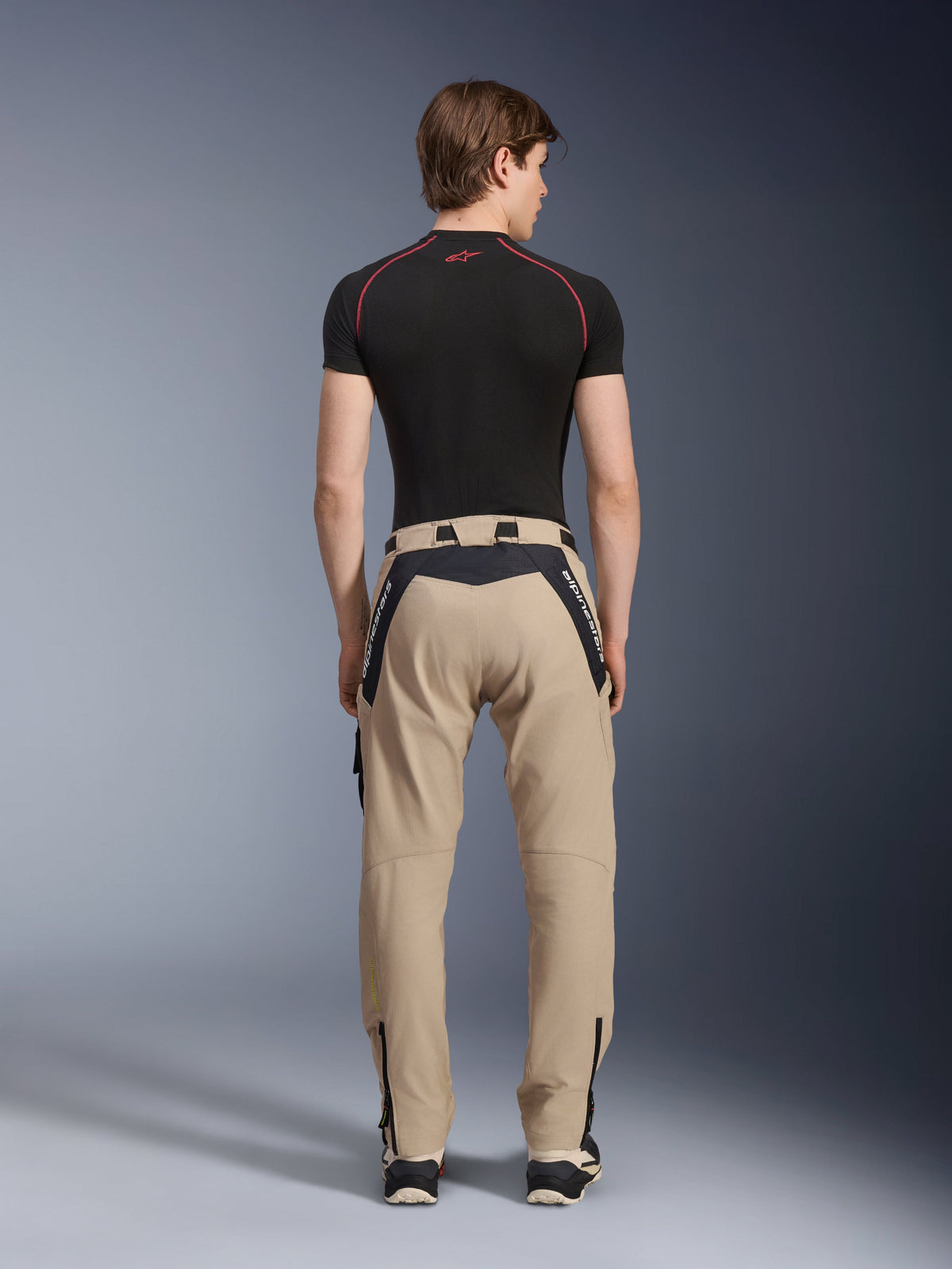 Alpinestars Acteon Pants in Sand, modern urban motorcycle pants viewed from the back, featuring black panels with white Alpinestars branding, tapered fit with zippers at the hem, worn with a black short-sleeved base layer and sneakers.