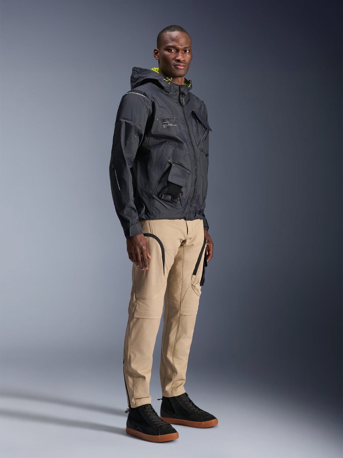 Alpinestars Acteon Pants, urban motorcycle pants, Sand color, worn by model with a dark technical jacket and black sneakers, featuring a tapered fit with black zipper accents and cargo pockets, contemporary motorsport-inspired design