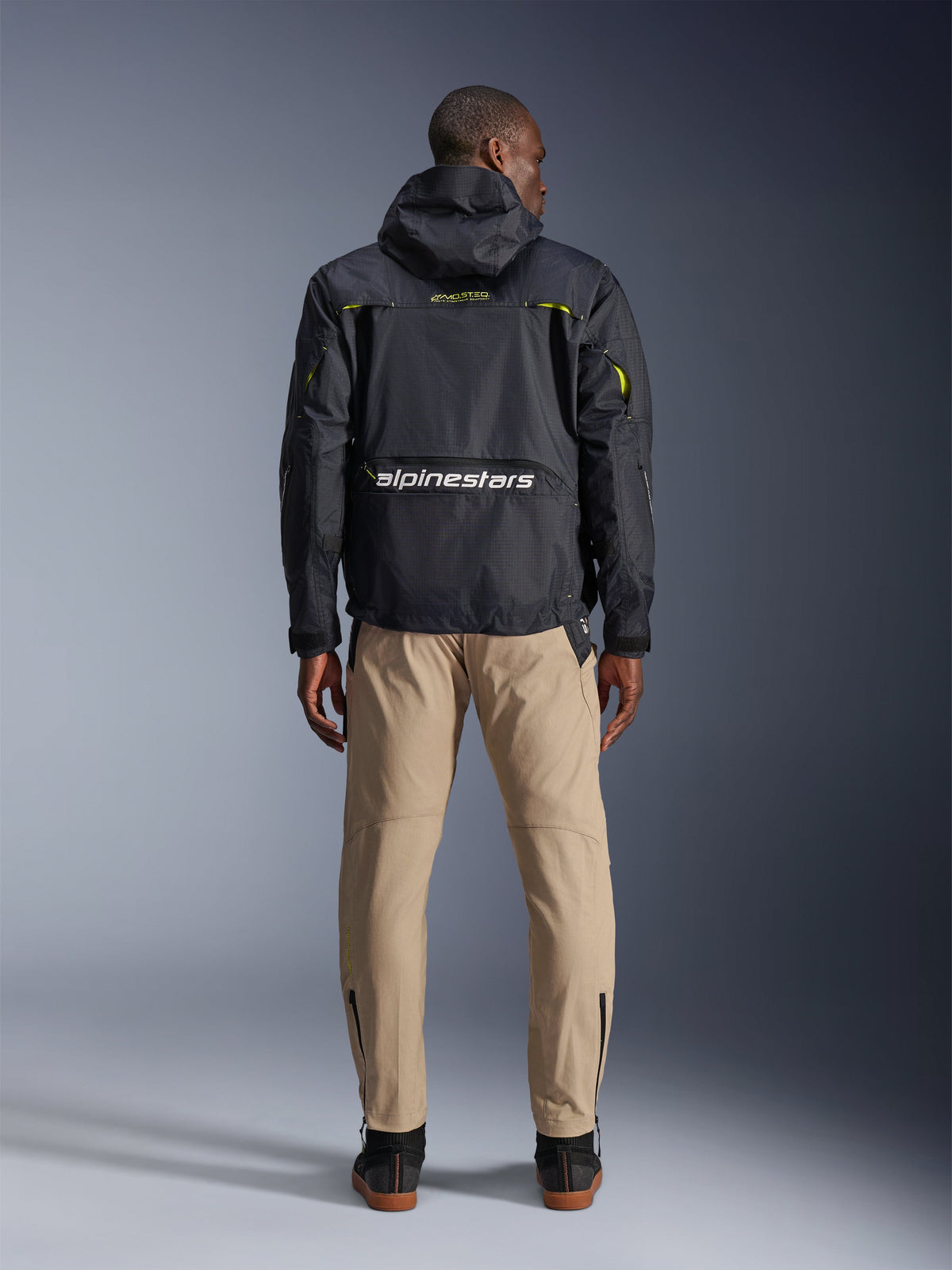 Alpinestars Acteon Pants, Urban Motorcycle Pants, Sand, shown from the back on a model wearing a black hooded technical jacket and dark boots, featuring a tapered leg with ankle zippers and a clean, modern silhouette.