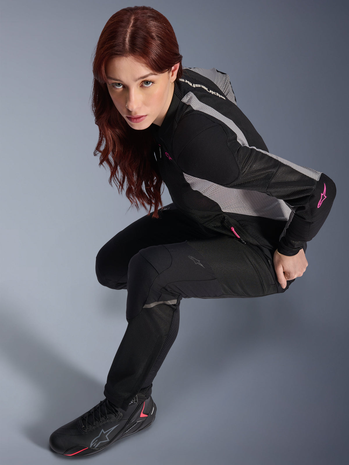 Woman wearing Alpinestars Stella Troop-Air Pants, Black, lightweight ventilated textile pants, paired with a black and grey mesh jacket with pink accents and black riding boots, studio portrait, urban motorcycle riding gear