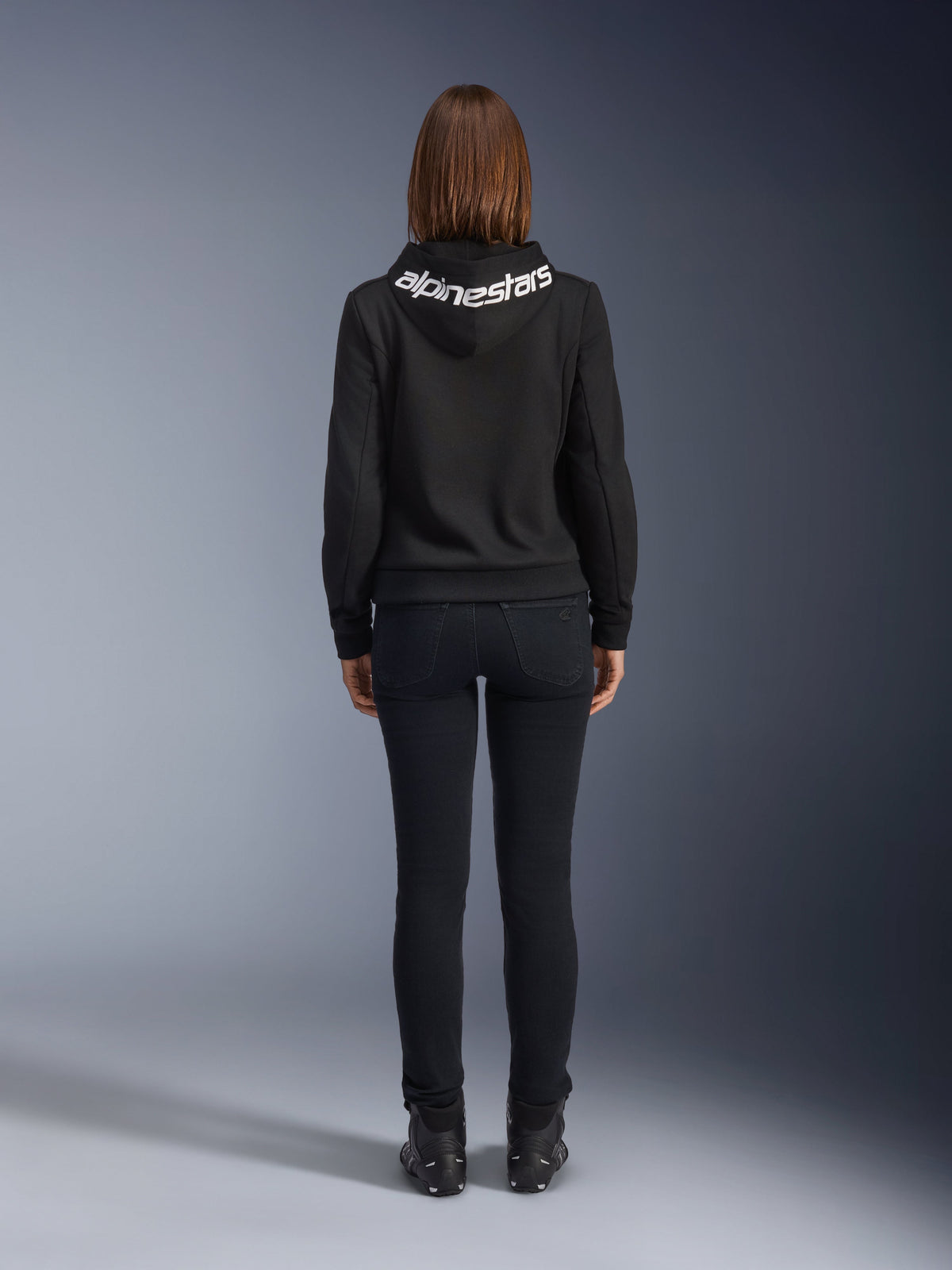 Daisy V3 Women's Riding Denim, Black Rinse, slim fit motorcycle pants, rear view showing high back waist and feminine silhouette, paired with a black Alpinestars hoodie and black riding boots, urban casual riding gear