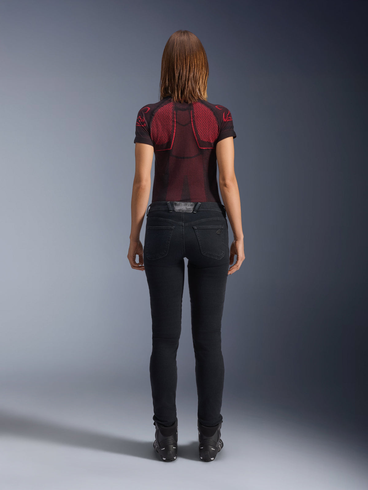 Alpinestars Daisy V3 Women's Riding Denim, Rinse Black, slim fit motorcycle pants shown from behind, featuring a high-waist design and back pockets, paired with a red and black patterned technical shirt and black boots, urban motorcycle gear for women