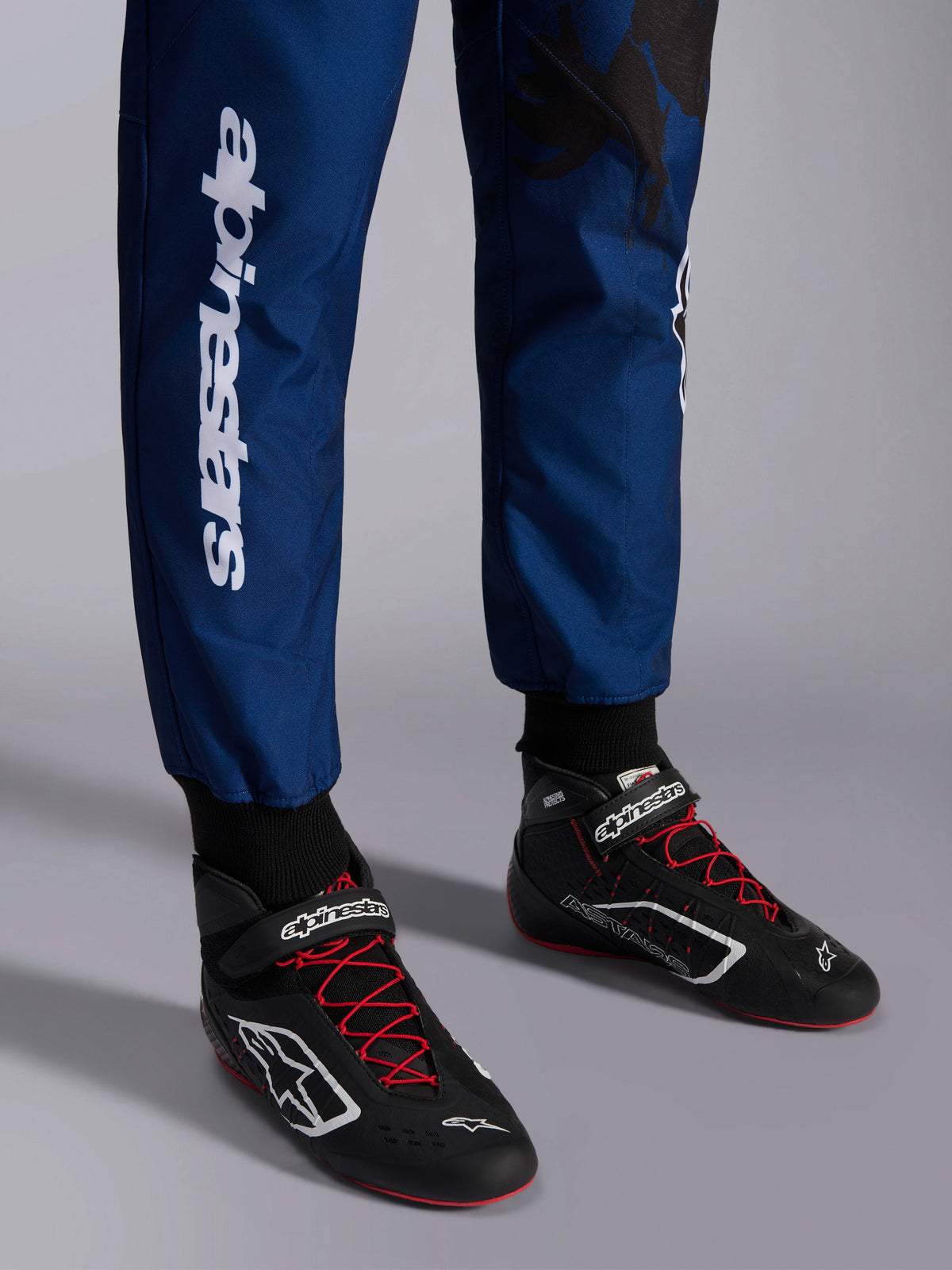 Alpinestars KMX-9 V3 Graphic 5 Suit, Navy Red, close-up of lower legs and karting shoes, navy blue fabric with white vertical Alpinestars branding, black shoes with red laces and white logos, professional karting gear