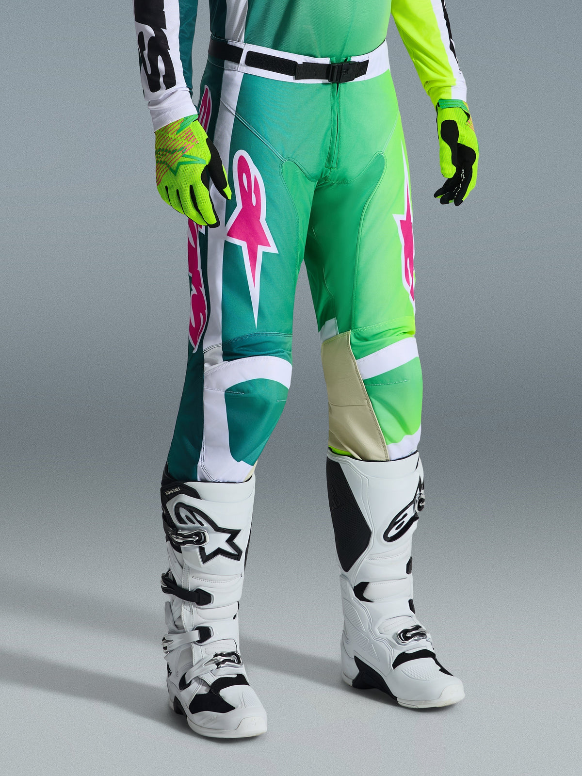 Alpinestars 2026 Racer Portl Pants, Motocross Pants, Green White and Black with pink logos, worn with white boots and neon yellow gloves, featuring bold graphic patterns and an adjustable black waist buckle, performance gear for off-road riding.