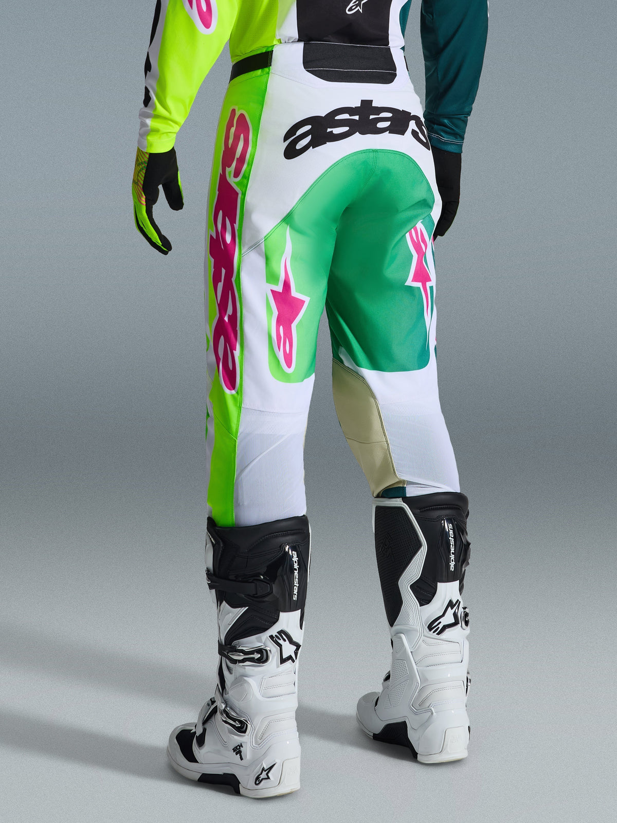 Alpinestars 2026 Racer Portl Pants, Motocross Pants, Green White and Black with pink accents, back view featuring bold 'astars' logo on the seat and pink star logos on legs, paired with white and black motocross boots, performance gear for off-road racing