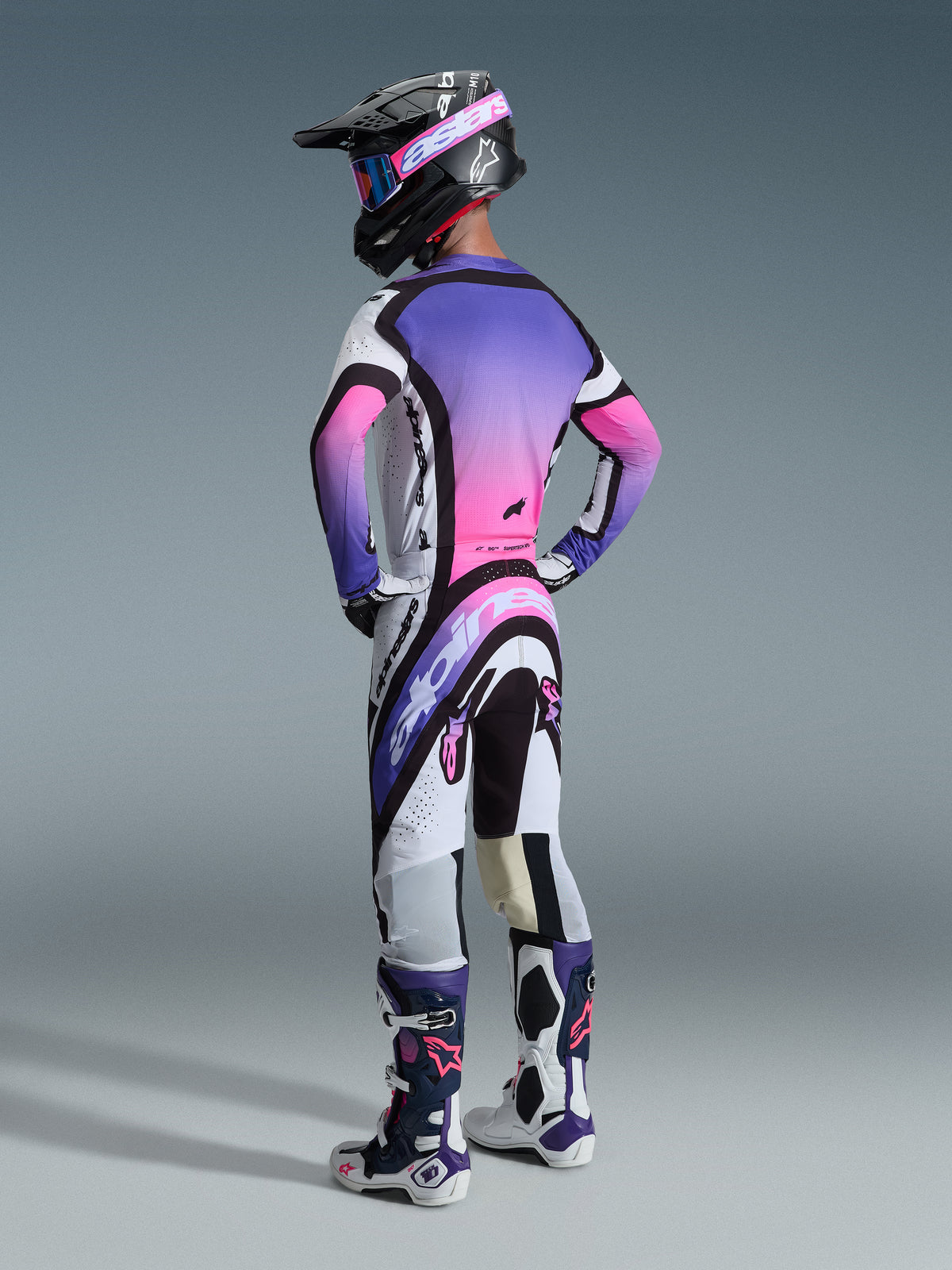 Motocross rider from behind wearing Alpinestars 2026 Supertech AFD Vista Pants, White Purple Black colorway, paired with a matching purple and pink gradient jersey, black helmet, and white and purple racing boots for a complete off-road racing look.