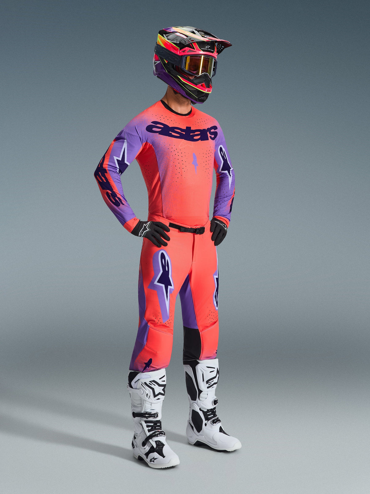 Motocross rider wearing full racing gear, featuring the Alpinestars 2026 Supertech Scenz Pants in Hot Coral Purple, matching coral and purple jersey, black and pink helmet, white and black boots, professional off-road racing kit.