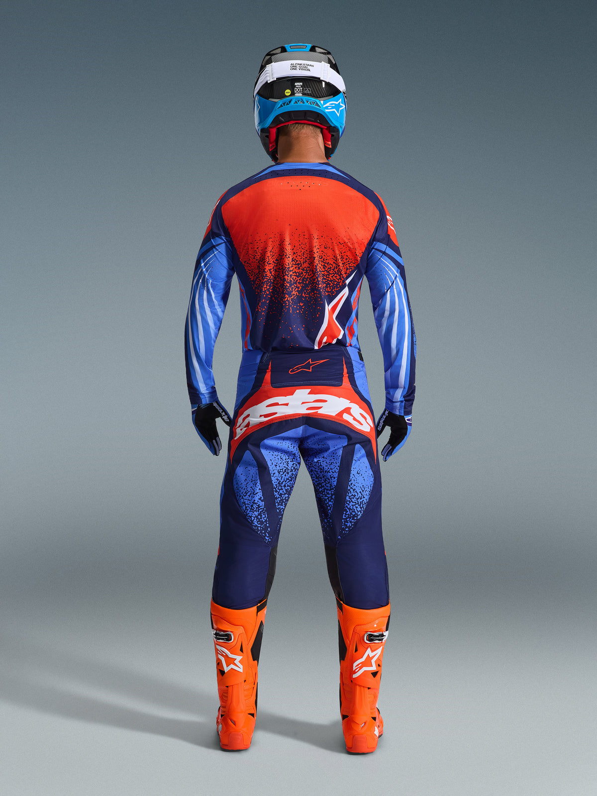 Motocross rider seen from the back wearing Alpinestars 2026 Techstar Nomur Pants in Dark Navy, Orange, and UCLA Blue, styled with a matching jersey and bright orange boots, professional off-road racing gear set.