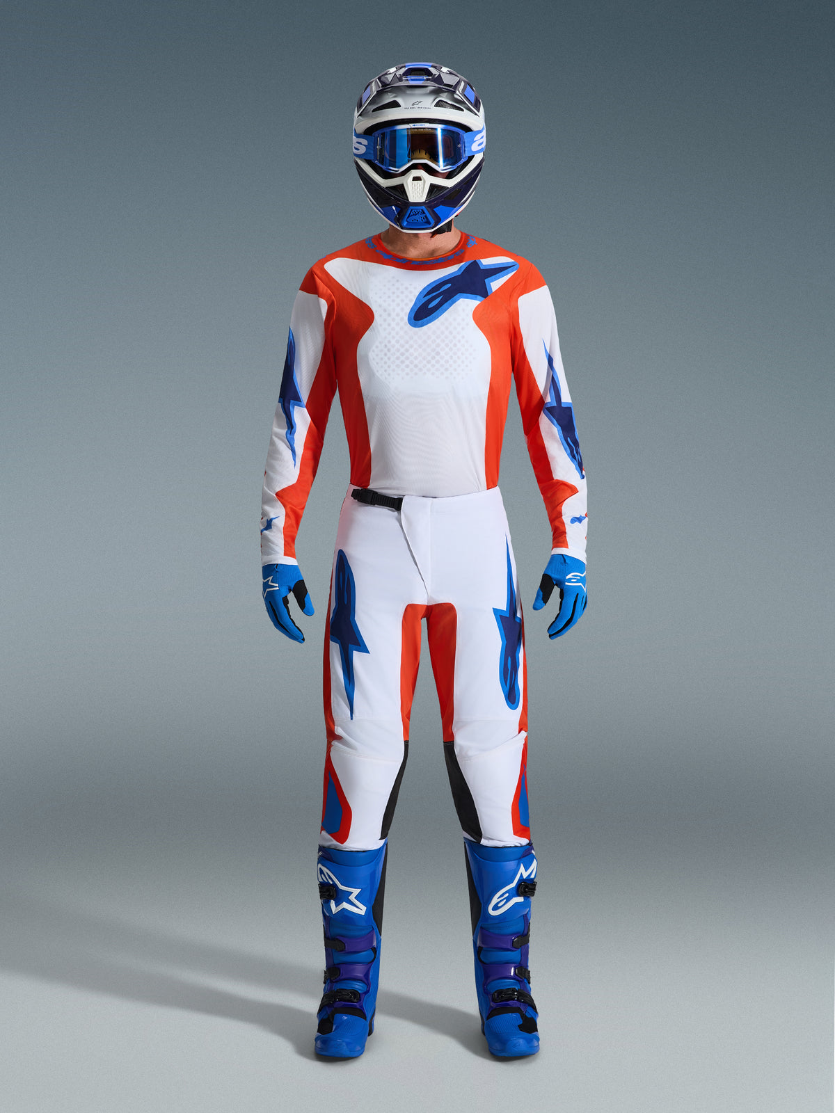 Motocross rider wearing complete racing gear, Alpinestars 2026 Fluid Grid Pants in Orange and UCLA Blue, white jersey with orange sleeves and blue logos, matching blue helmet and goggles, blue boots and blue gloves, coordinated outfit for off-road racing