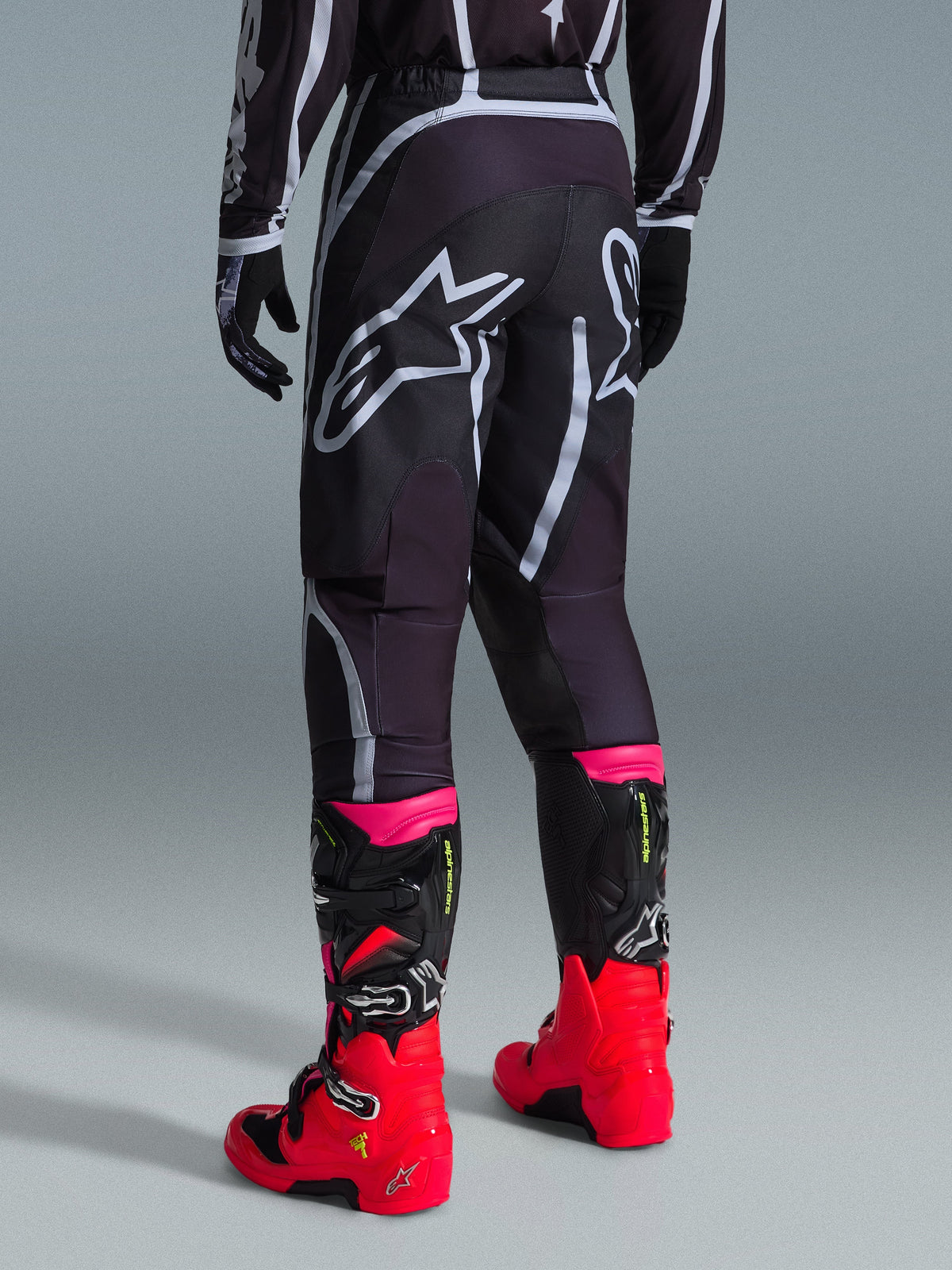 Alpinestars 2026 Fluid Apex Pants, Motocross Pants, Black and Gray, rear view featuring large white Alpinestars logos on the seat, worn with bright red and pink boots, durable 600D polyfabric construction for off-road performance