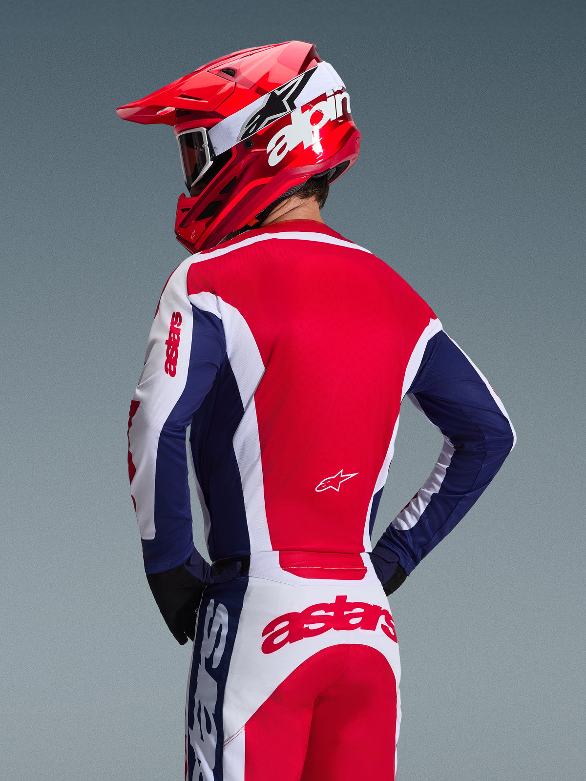 Motocross rider from behind wearing Alpinestars 2026 Racer Riway Jersey, White Blue and Red colorway, matching off-road pants with bold astars branding, red helmet with white Alpinestars logo, black gloves, coordinated motocross racing gear