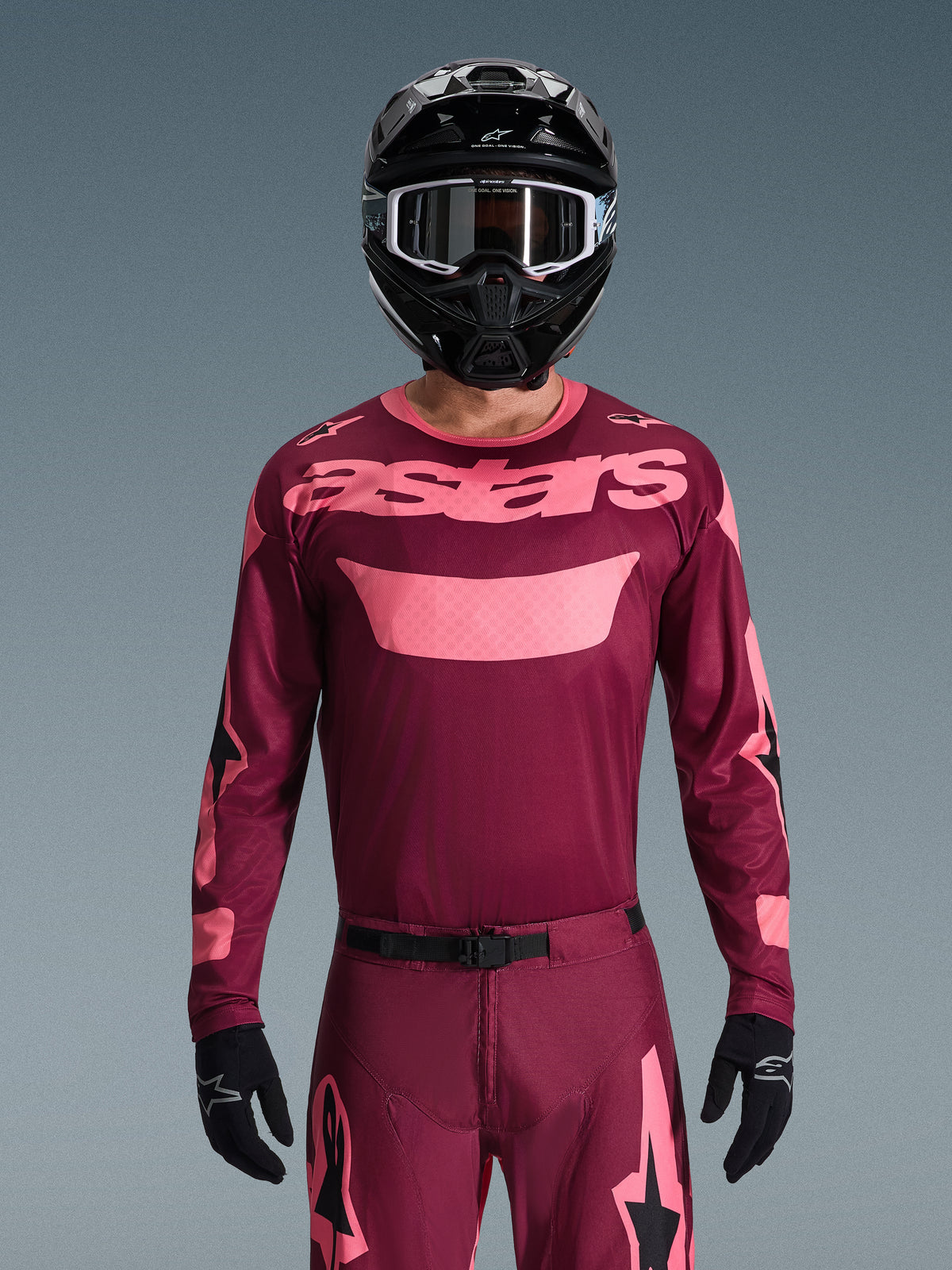 Motocross rider wearing Alpinestars 2026 Racer Riway Gear Set, Maroon and Pink, jersey with bold pink branding on the chest, matching pants with black and pink accents, black full-face helmet with goggles, and black gloves, coordinated off-road racing outfit.