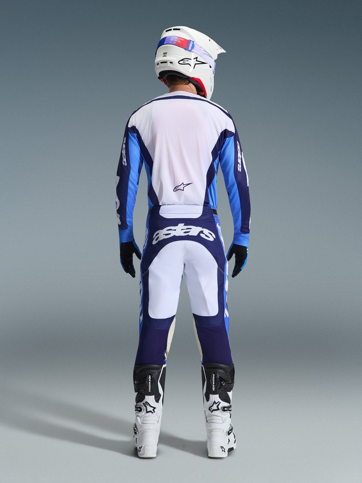 Motocross rider from behind wearing the Alpinestars 2026 Racer Riway Jersey in Ucla Blue and White, matching motocross pants with white and navy blue panels and Astars branding, white and black boots, and a white helmet, complete coordinated racing outfit for off-road performance.