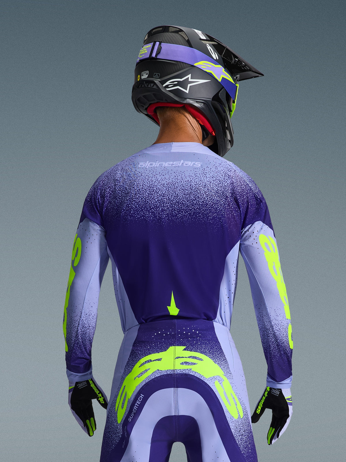 Alpinestars 2026 Supertech Scenz Jersey in Purple and Light Blue with Fluorescent Yellow accents, back view on rider wearing matching motocross pants, black helmet, and gloves, high-performance athletic gear for off-road racing