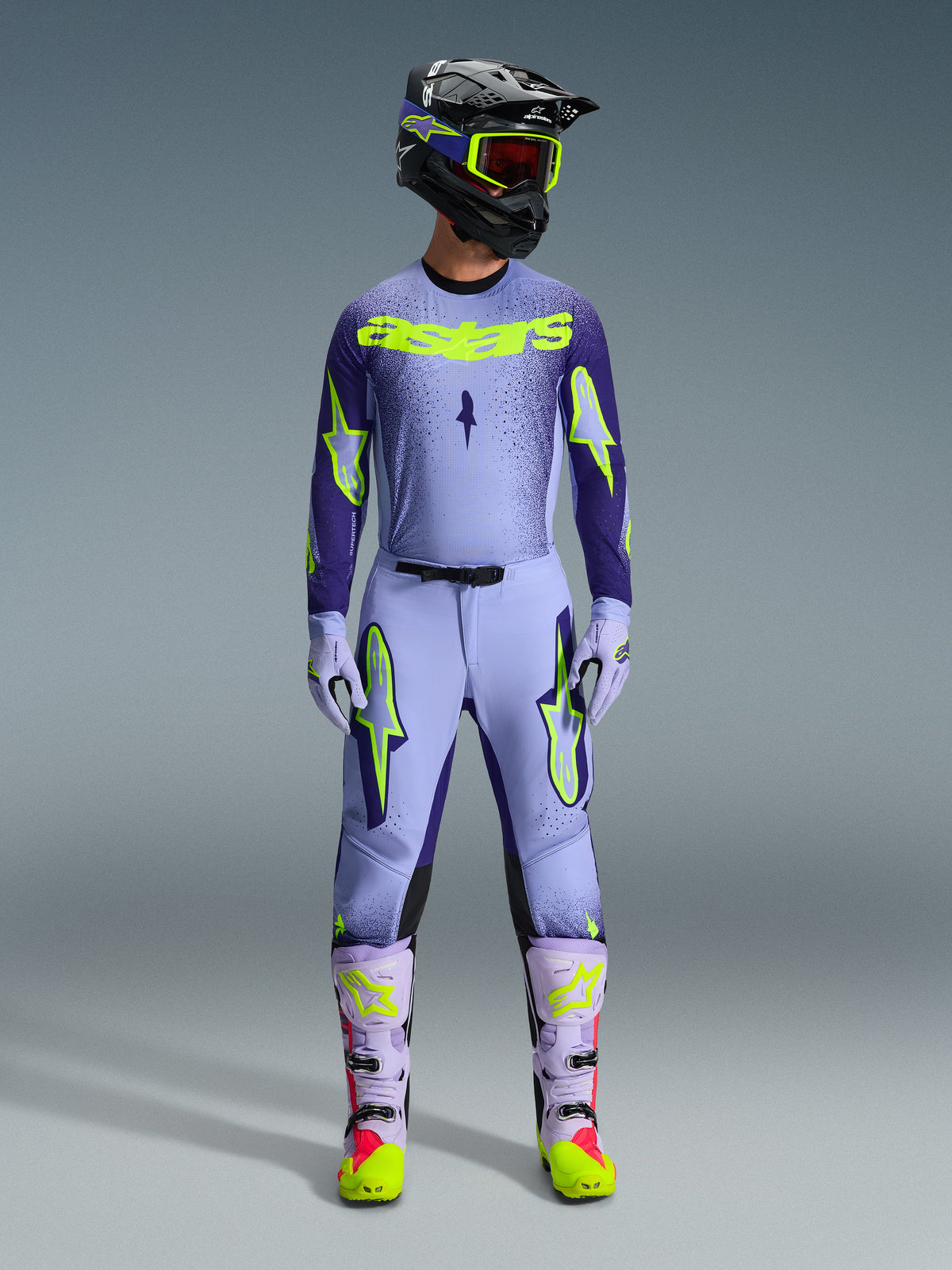 Motocross rider wearing complete Alpinestars 2026 Supertech Scenz racing gear, Purple and Yellow Fluo jersey with matching pants, black helmet with yellow goggles, light purple and neon boots, professional athletic-fit outfit for off-road racing