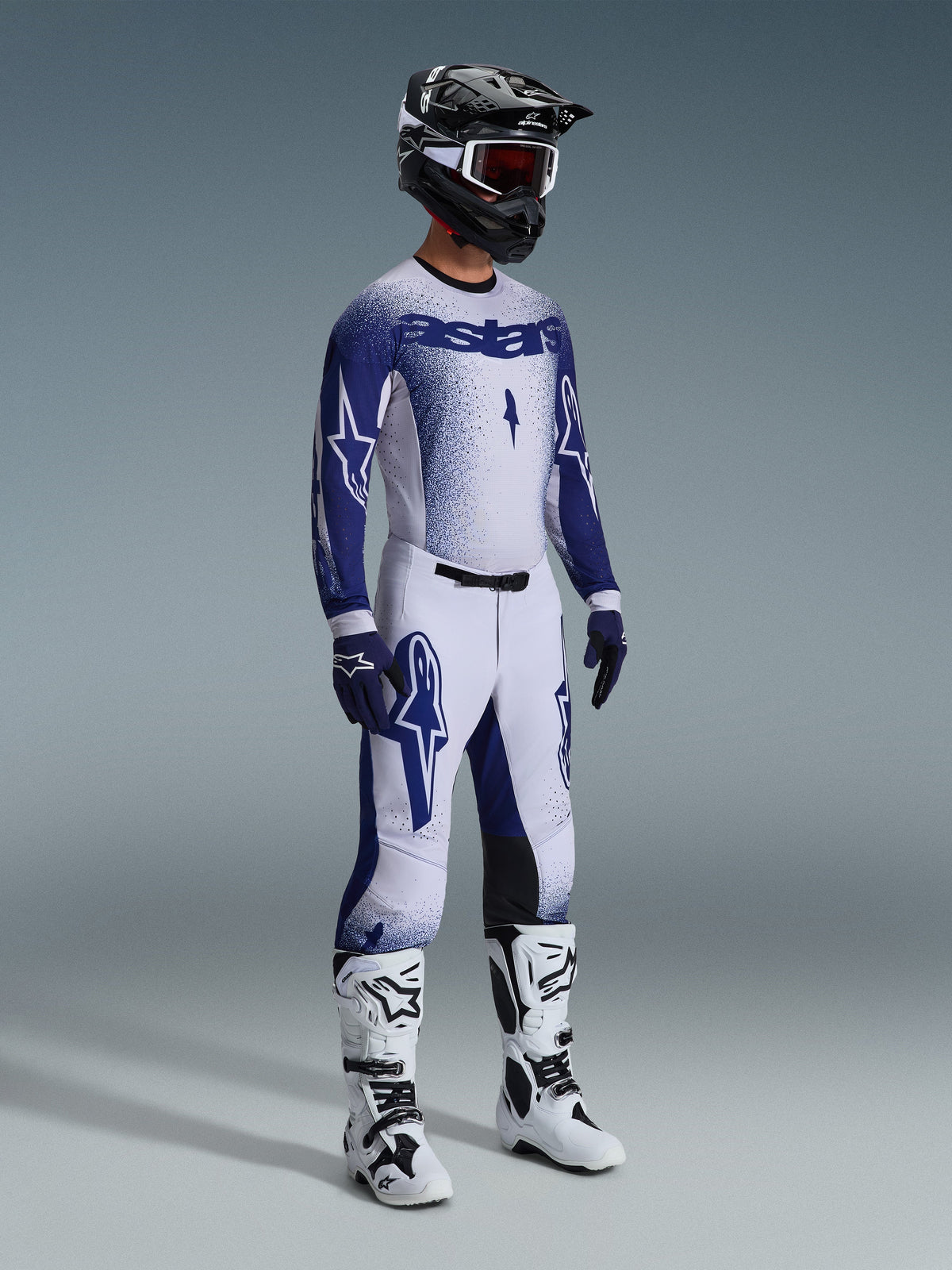 Motocross rider wearing complete professional racing gear, 2026 Supertech Scenz Jersey in light gray and dark blue, matching motocross pants with star graphics, black helmet with red goggles, white and black performance boots, coordinated blue and white gloves for off-road racing