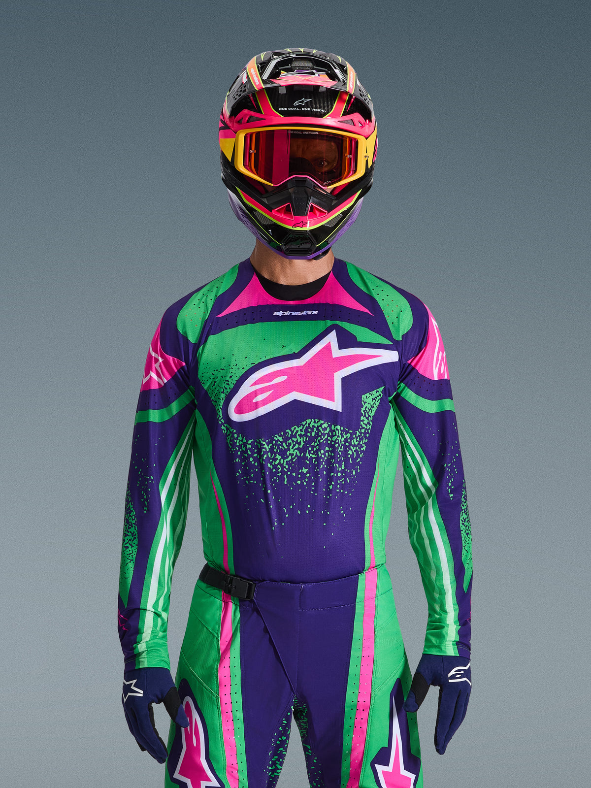 Alpinestars 2026 Techstar Nomur Gear Set, Motocross rider wearing vibrant green and purple racing jersey with pink accents, matching gear set with multi-colored helmet and goggles, high-performance off-road motorcycle outfit