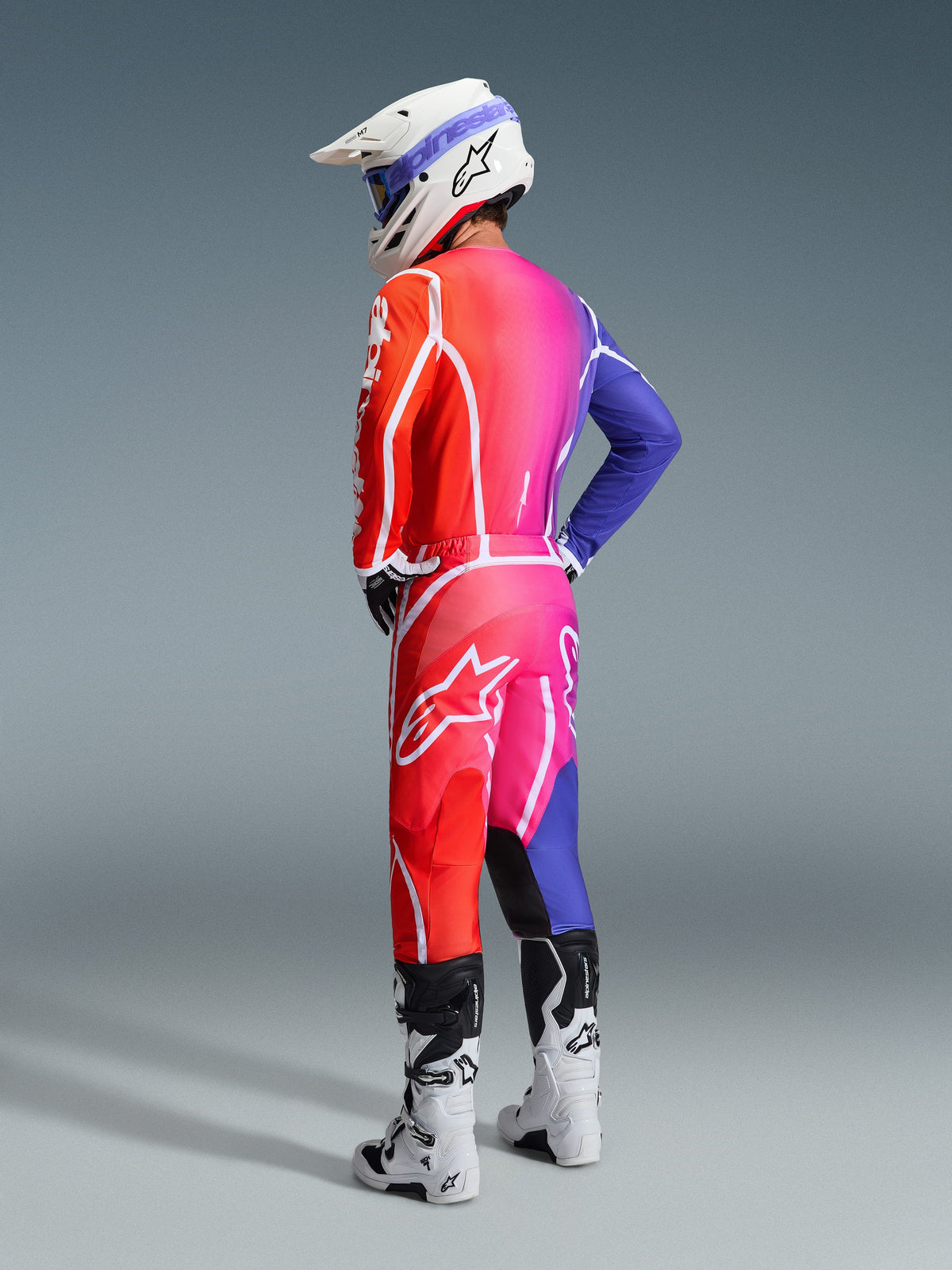 Motocross rider wearing the Alpinestars 2026 Fluid Apex Jersey and matching pants, vibrant red, pink, and blue gradient with white accents, white helmet with blue goggles and white boots, rear view of complete off-road racing gear.
