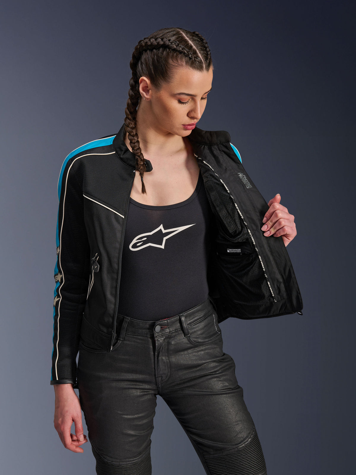 Woman wearing the Alpinestars Stella T-Dyno Air Jacket in Black Light Blue Ecru, holding the jacket open to reveal the inner mesh lining and waterproof pocket, paired with a black logo tank top and black riding pants, vintage-inspired urban motorcycle gear.