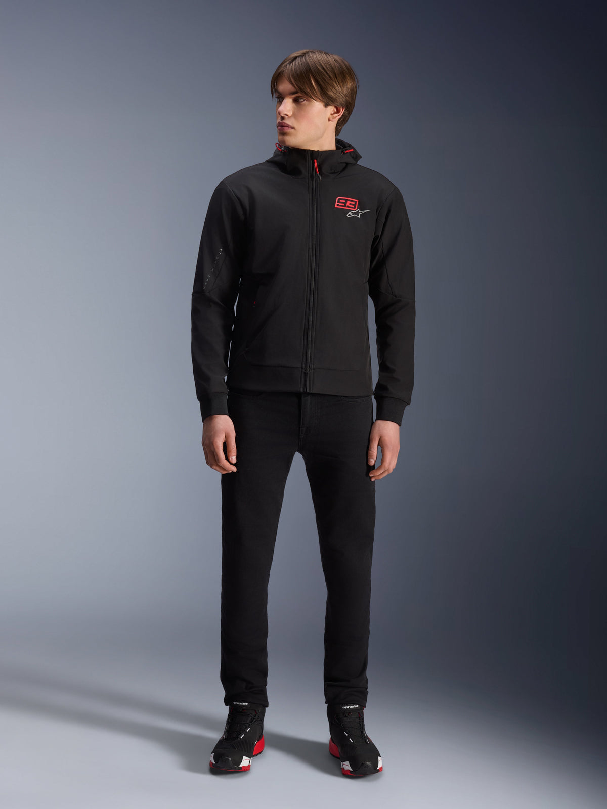 Alpinestars MM93 Radium Tech Hoodie, black urban motorcycle hoodie, featuring a red 93 logo and white star logo on the chest, worn by a model with black pants and matching riding shoes, sleek windproof technical shell for urban riding.
