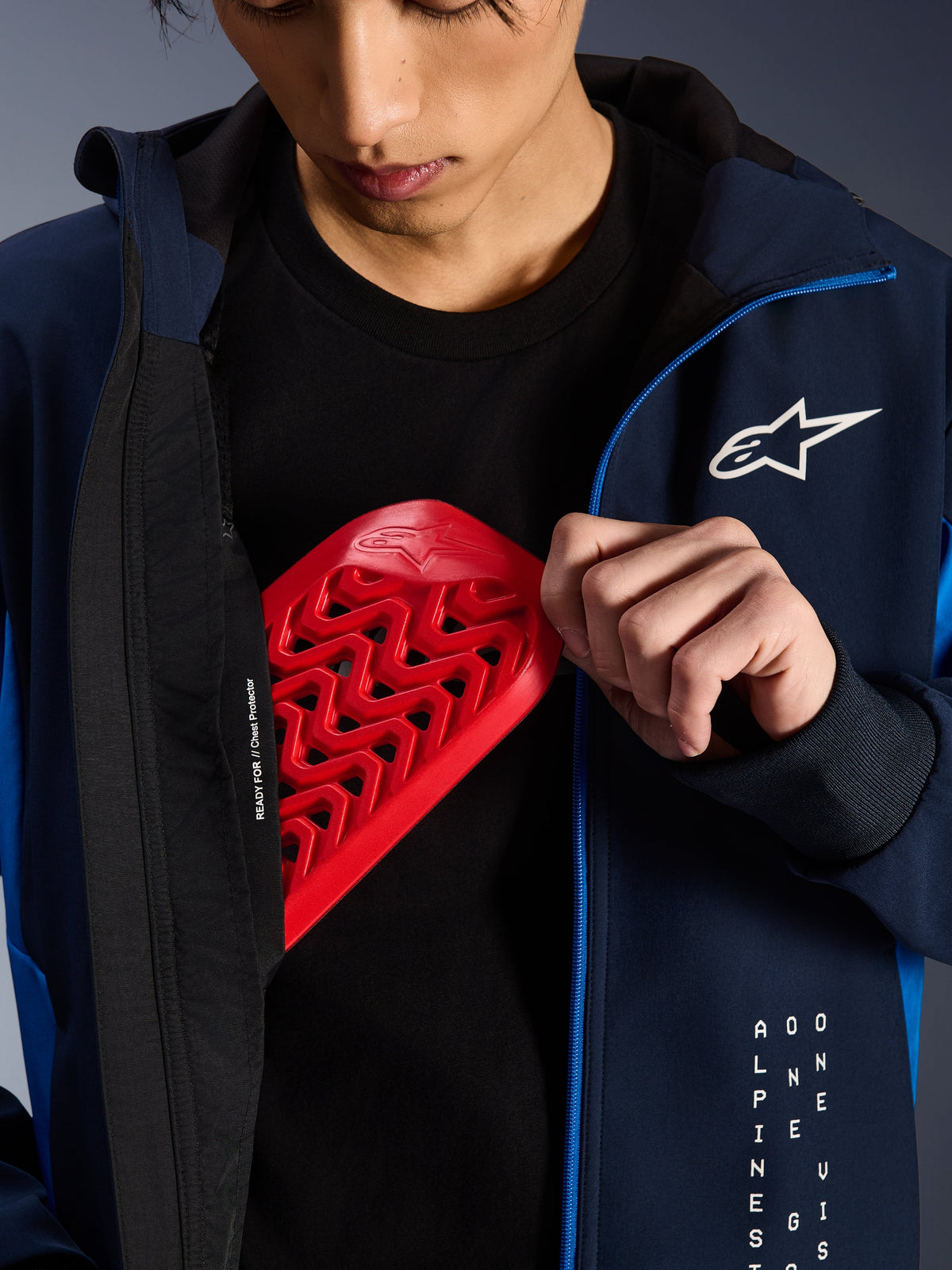 Alpinestars Radium Tech Hoodie in Night Navy and Royal Blue, close-up showing a red Nucleon chest protector being placed into the internal pocket, featuring 'READY FOR // Chest Protector' text on the lining and a white star logo on the chest.