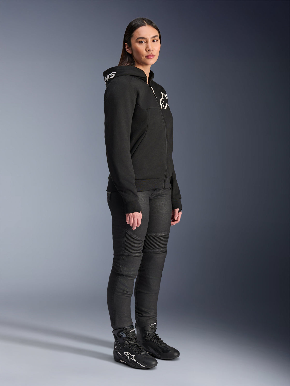 Woman wearing Alpinestars Stella Chrome V2 Sport Hoodie in black, featuring white Alpinestars branding, paired with black motorcycle riding pants and boots, urban riding gear for women with a casual athletic fit