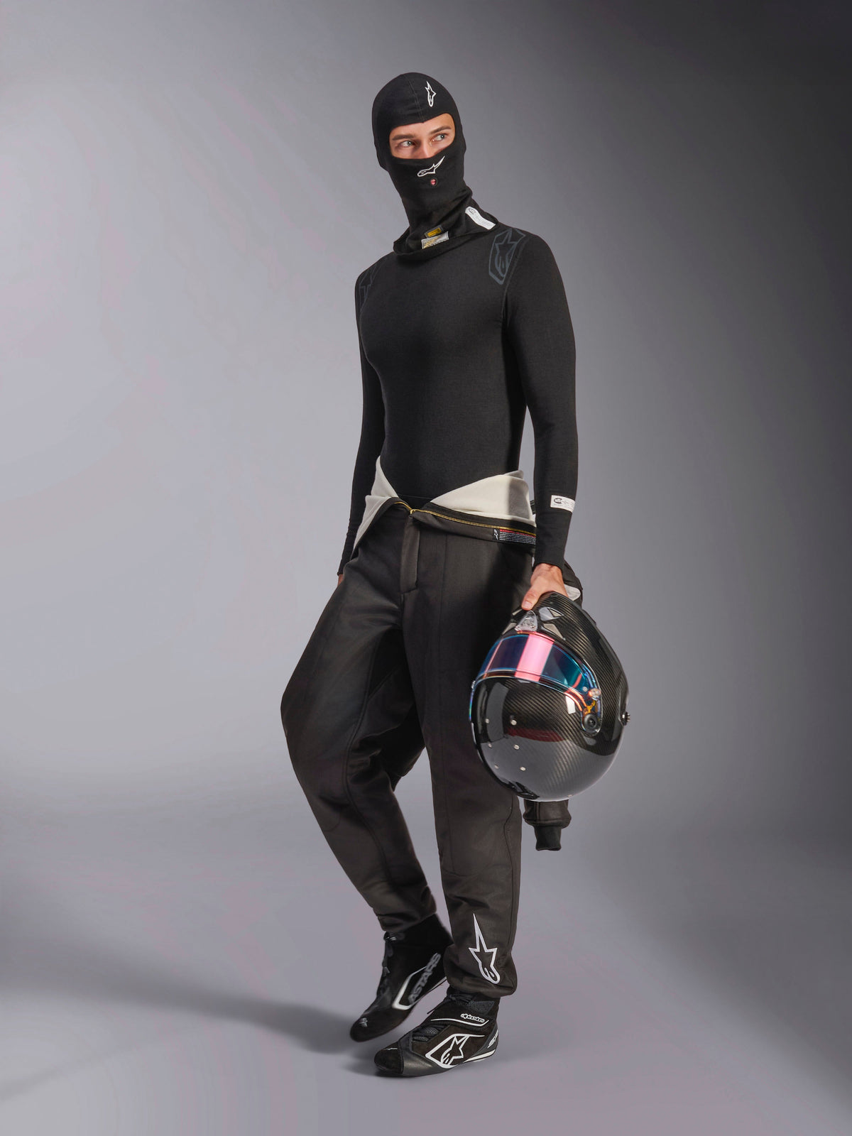 Alpinestars ZX Evo V3 Balaclava, Black, worn by a driver with complete auto racing base layer gear, featuring the white logo on the forehead, holding a carbon fiber helmet with an iridescent visor, professional fire-resistant motorsport apparel.