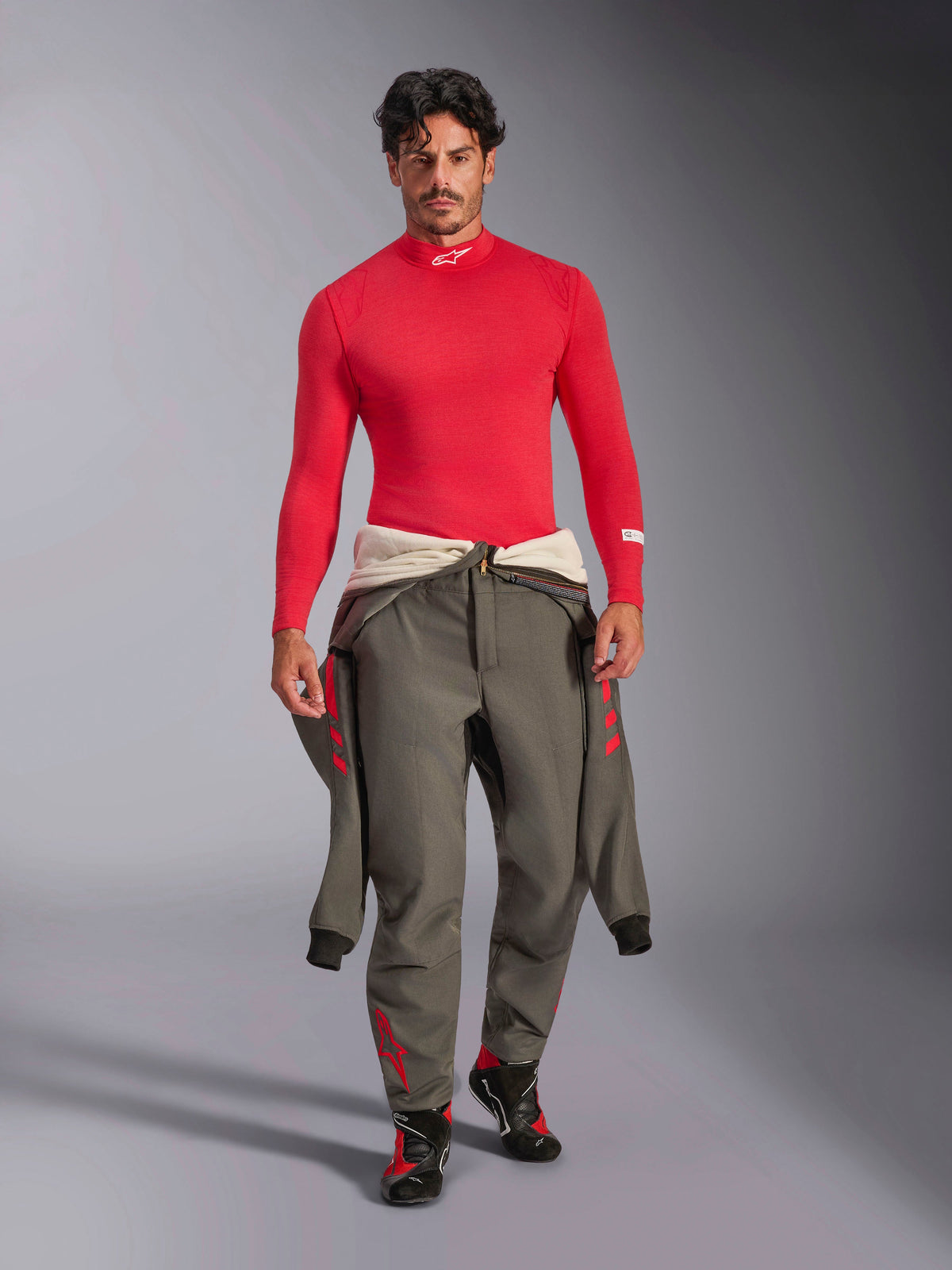 Alpinestars ZX Evo V3 Long Sleeve Top, Red Auto Base Layer, worn by a model with a grey racing suit tied at the waist and racing shoes, mock neck with white Alpinestars logo, lightweight and flame-resistant professional motorsport gear.