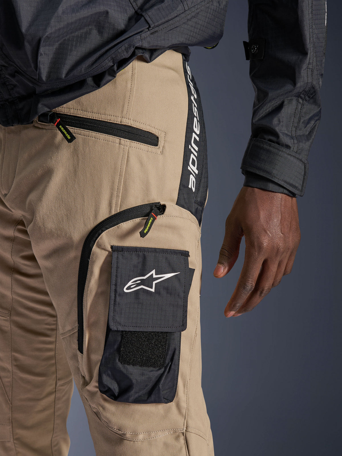 Alpinestars Acteon Pants in Sand, detailed view of the hip and thigh area, featuring a black side cargo pocket with white star logo, zippered pockets, and vertical Alpinestars branding on a black side panel, urban motorcycle apparel.