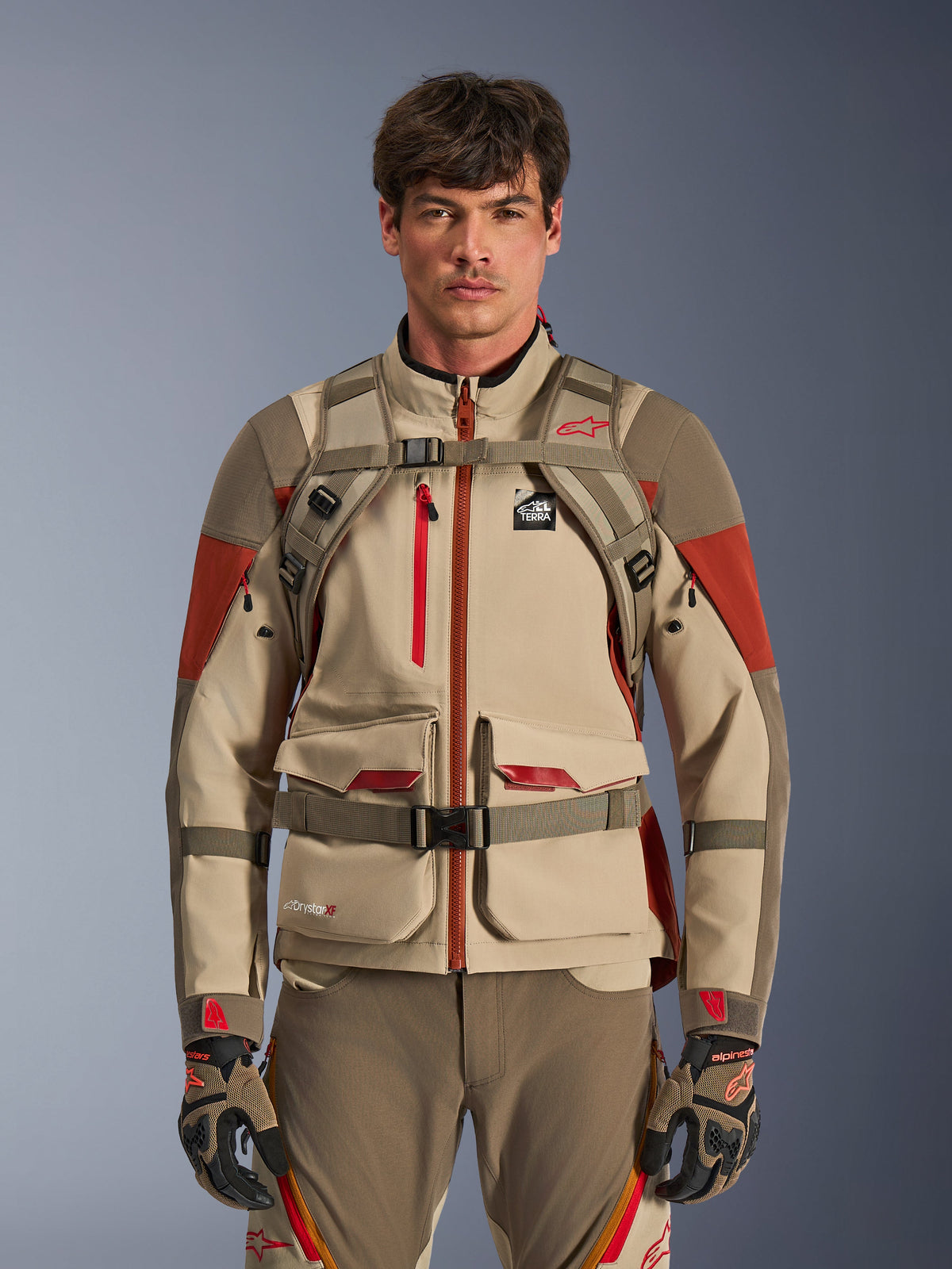 Adventure rider wearing Alpinestars AT-15 Backpack in Aluminum Walnut, front view showing chest and waist straps with magnetic lock, paired with tan and red adventure motorcycle jacket and pants, technical off-road gear ensemble