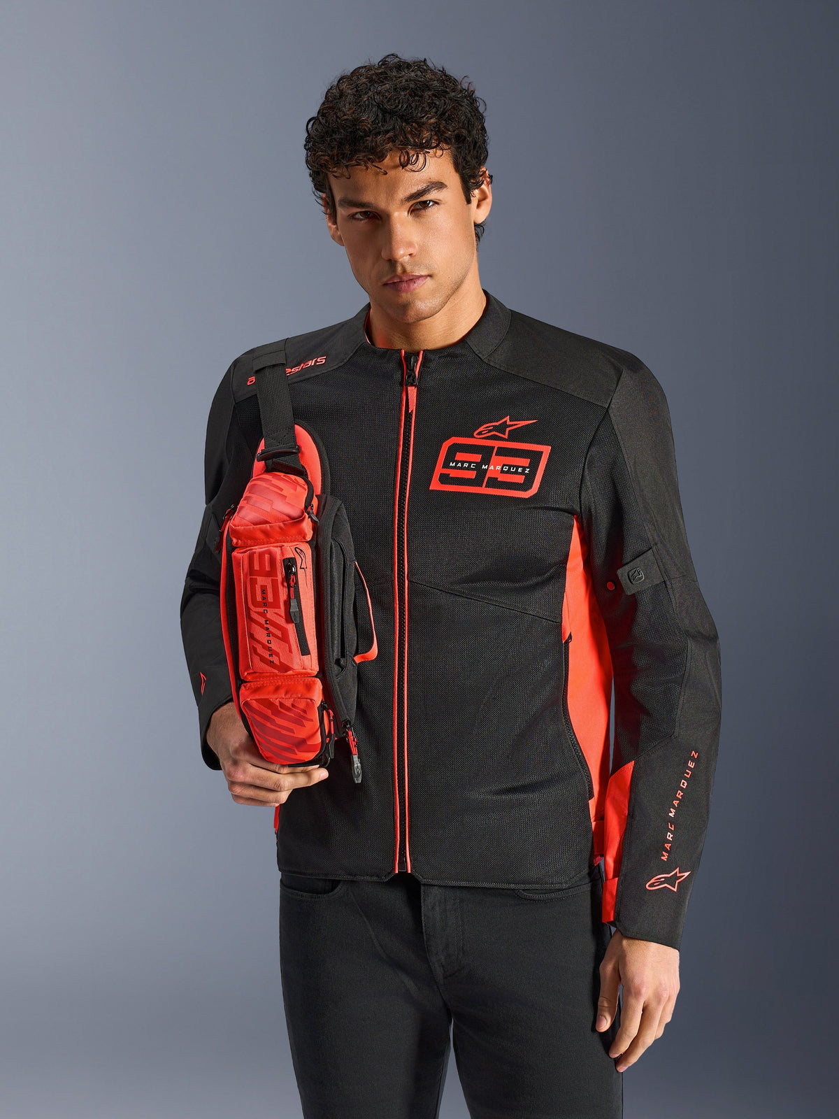 Man wearing Alpinestars MM93 Waist Bag, Red Fluo and Black, styled as a crossbody bag over a matching black and red motorcycle jacket, featuring Marc Marquez 93 logo, sporty racing design