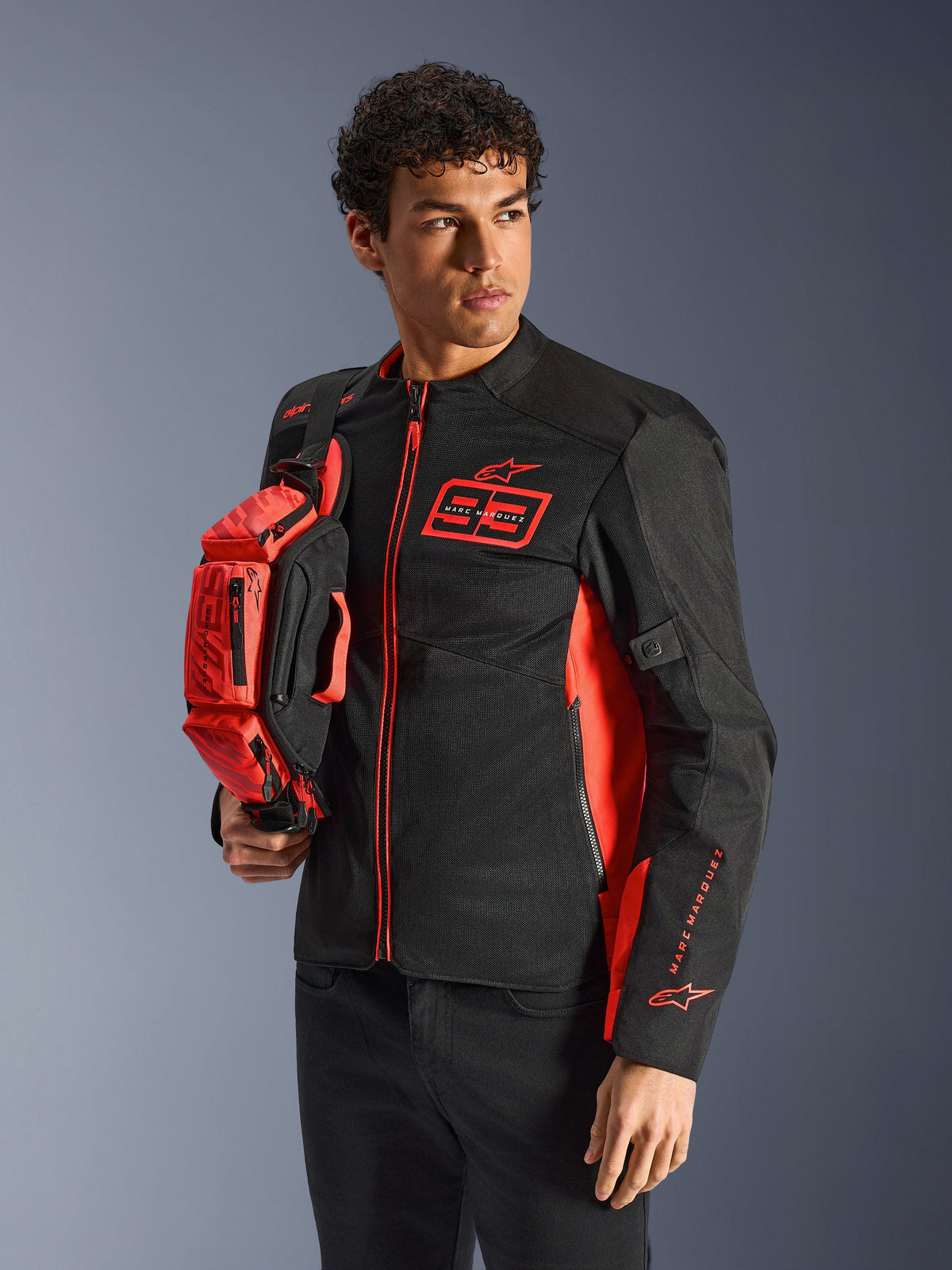 Alpinestars MM93 Waist Bag, Red Fluo and Black, held by a model wearing a matching black and red motorcycle jacket, featuring multiple zippered compartments and Marc Marquez branding, practical gear for sport riding