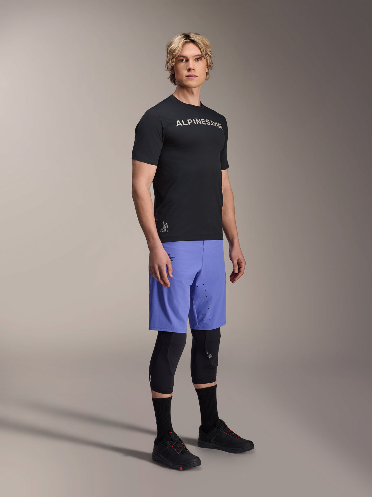 Male model wearing Alpinestars A-Aria Switch Short Sleeve Jersey in Black with white chest branding, paired with light blue mountain bike shorts, black knee guards, and black cycling shoes, studio full-body shot for performance MTB gear