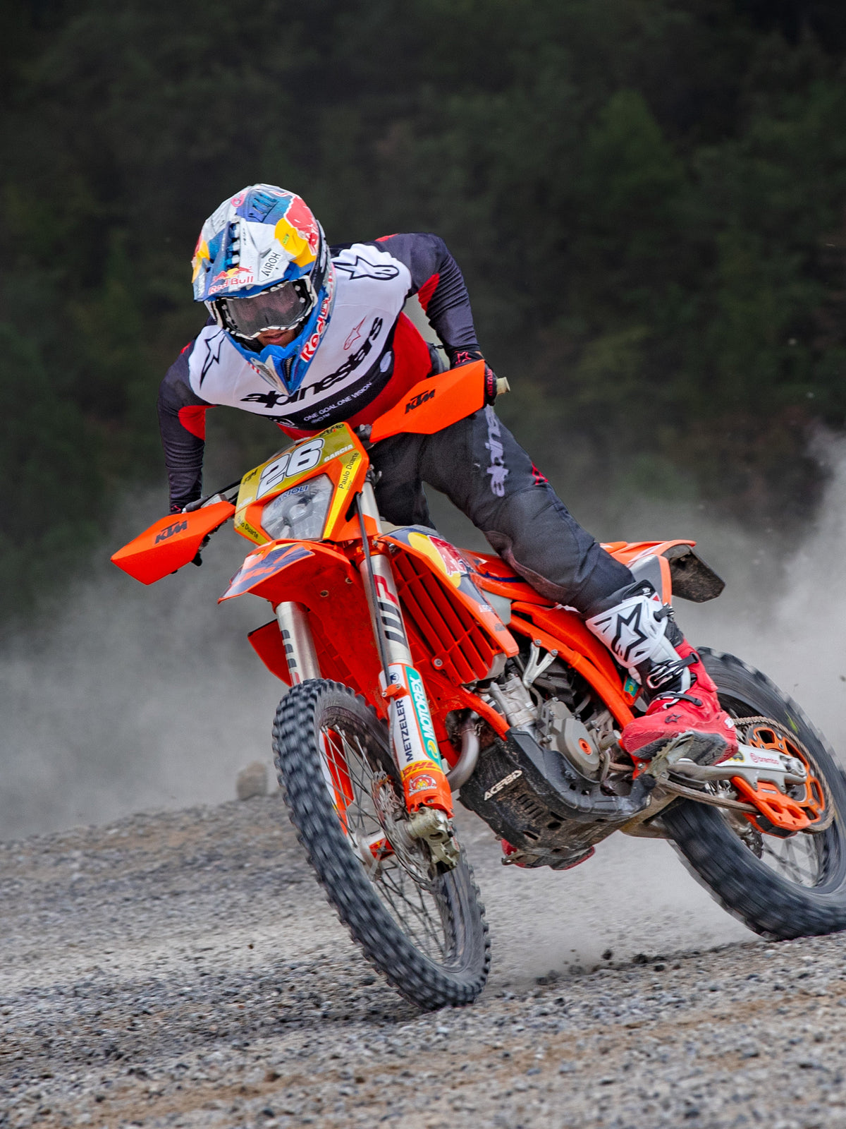 Motocross rider in action on an orange KTM bike, wearing Alpinestars Pro-Dura gear including a red, white, and black jersey and dark grey pants, red and white boots, and a Red Bull racing helmet, performing a sharp turn on a gravel trail
