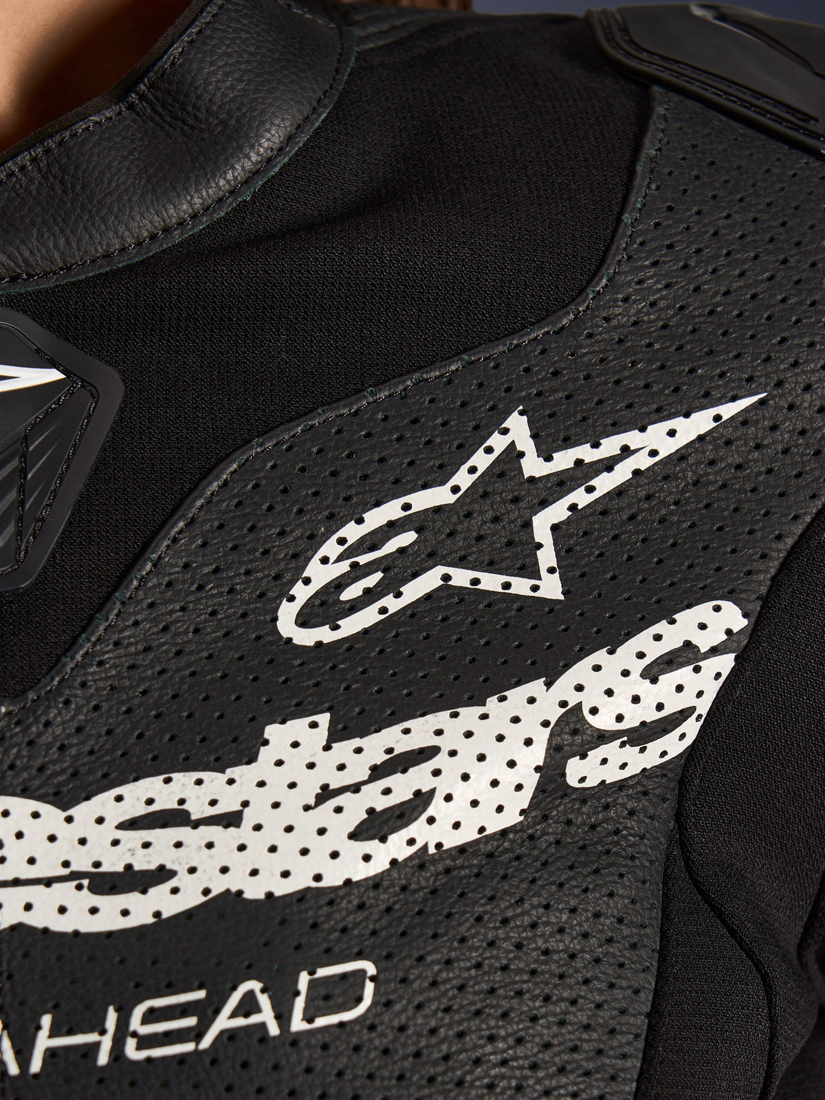 Close-up of the Alpinestars Stella GP-R7 2-Piece Leather Suit in Black, featuring perforated leather panels, a large white Alpinestars logo on the chest, and breathable stretch fabric sections.